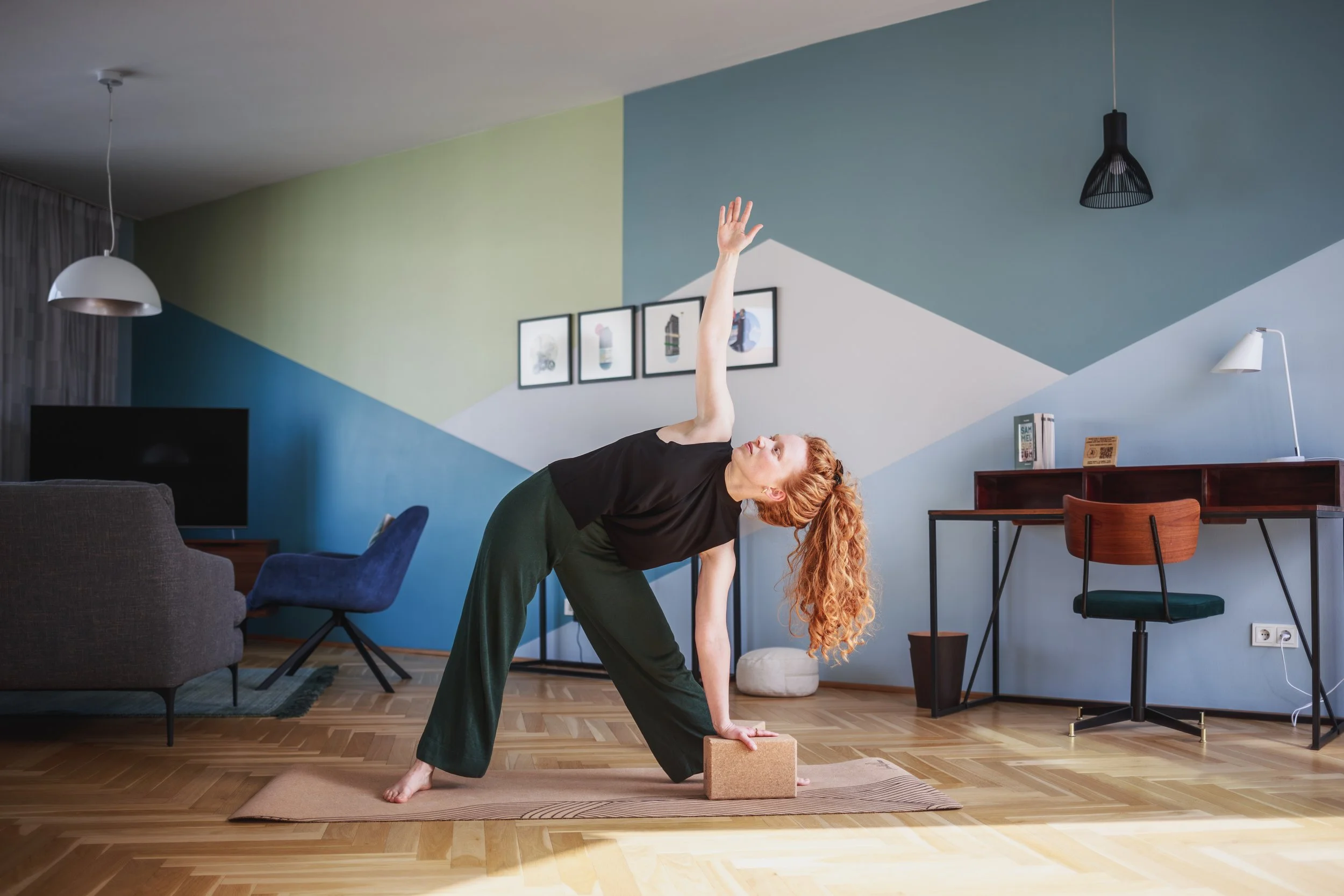 Eine Frau macht Yoga im Wohnzimmer, in der Haltung des seitlichen Dreiecks, die Hand nach oben gestreckt, auf einer Yoga-Matte. Das Zimmer hat bunte, geometrische Wandgestaltungen, Wandbilder, eine Stehlampe, einen Fernseher und einen Schreibtisch.
