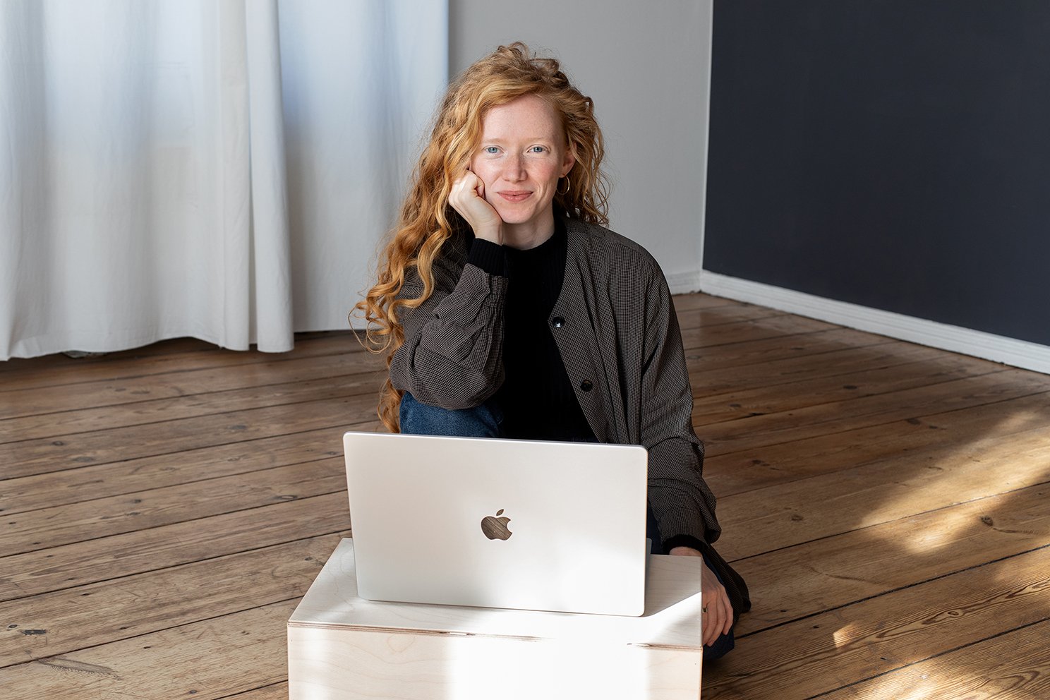 Eine Frau mit roten, lockigen Haaren sitzt auf dem Boden vor einem weißen Laptop.