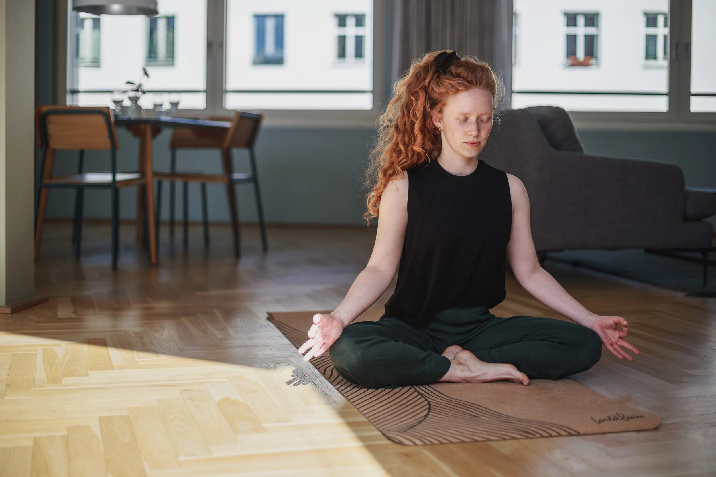 Eine Frau sitzt im Schneidersitz auf einem Yoga- oder Meditationsmatte in einem hellen Raum vor großen Fenstern, entspannt mit geschlossenen Augen, während sie meditiert oder Yoga praktiziert.