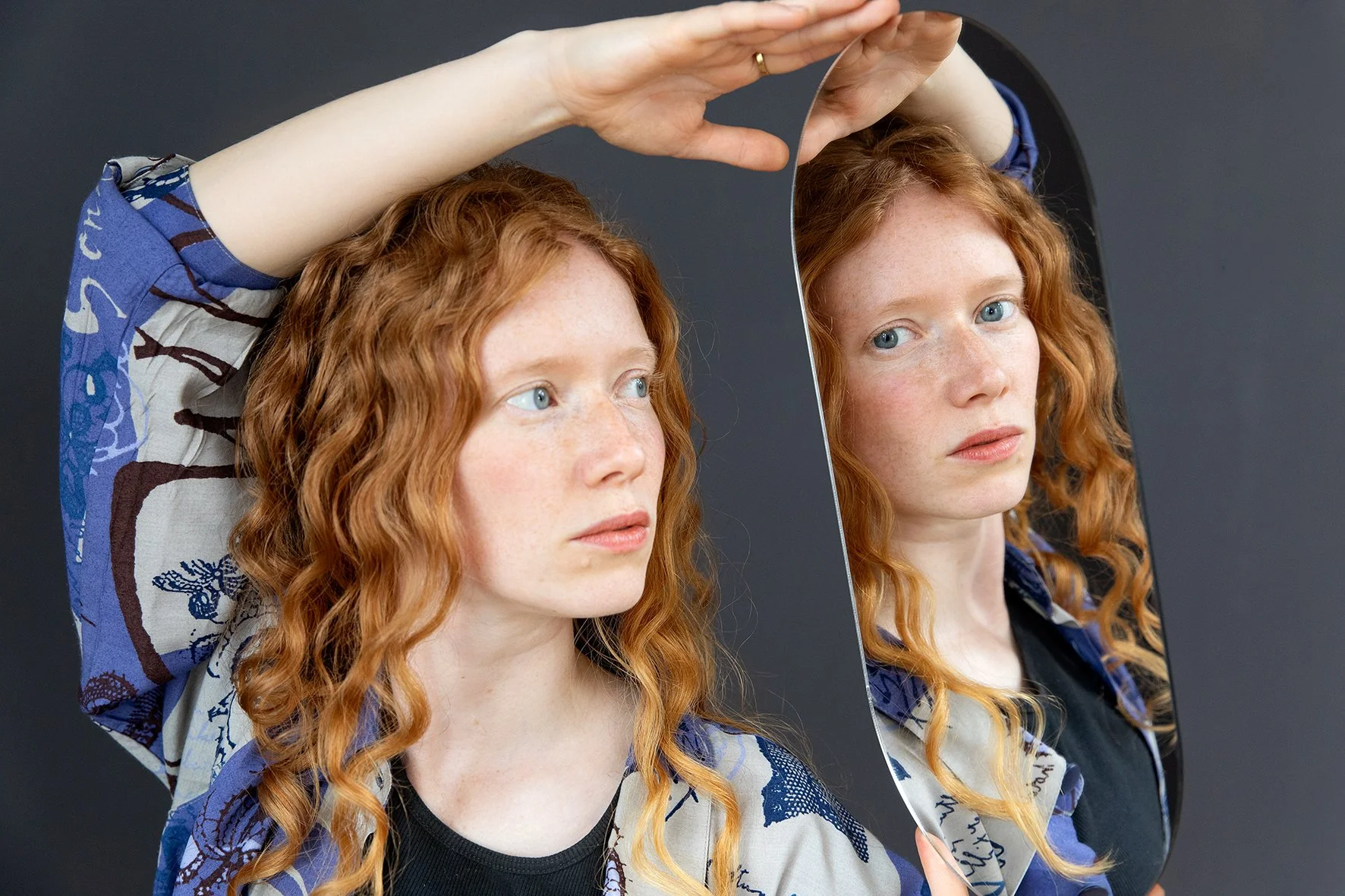 Eine rothaarige Frau schaut in einen kleinen Handspiegel, der ihren reflektiert. Sie trägt eine farbenfrohe Bluse und hat lockige Haare. Der Hintergrund ist dunkel, was die Person hervorhebt.