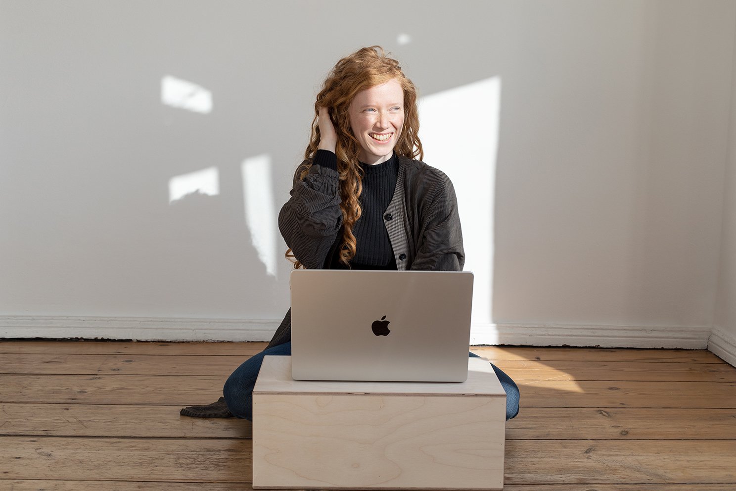 Eine junge Frau mit roten Locken sitzt auf dem Boden vor einem Laptop und lächelt, während sie in einem hellen Raum mit Holzboden ist.