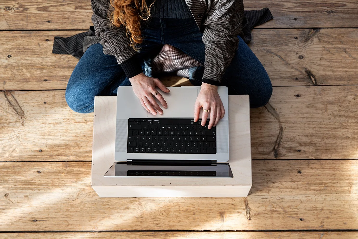 Person sitzt auf einem Holzpodest, arbeitet an einem Laptop, der auf einem offenen Kistentisch steht. Die Person trägt eine graue Jacke und blaue Jeans.