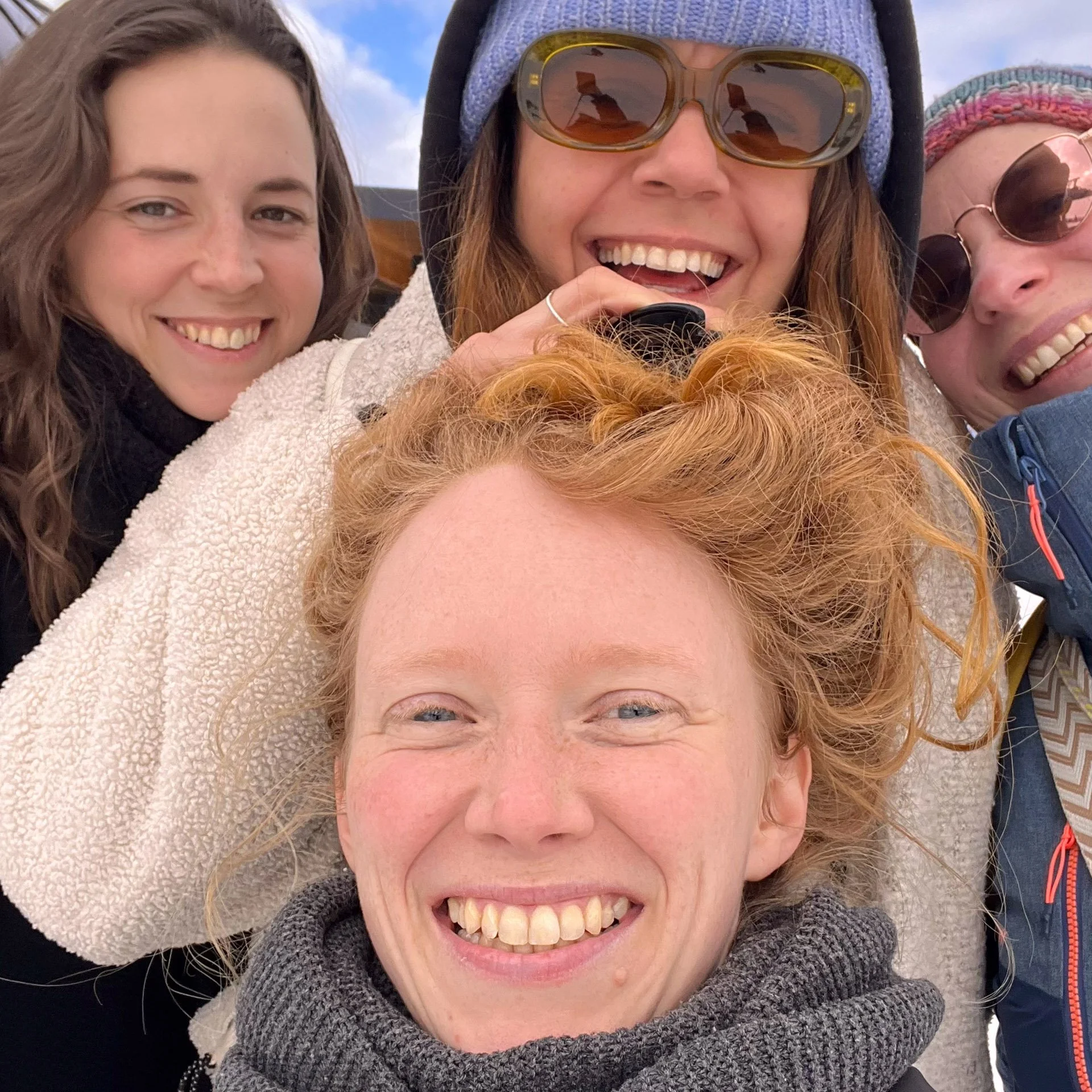 Drei lachende Frauen liegen eng aneinander, während sie ein Selfie machen, im Freien bei blauem Himmel.