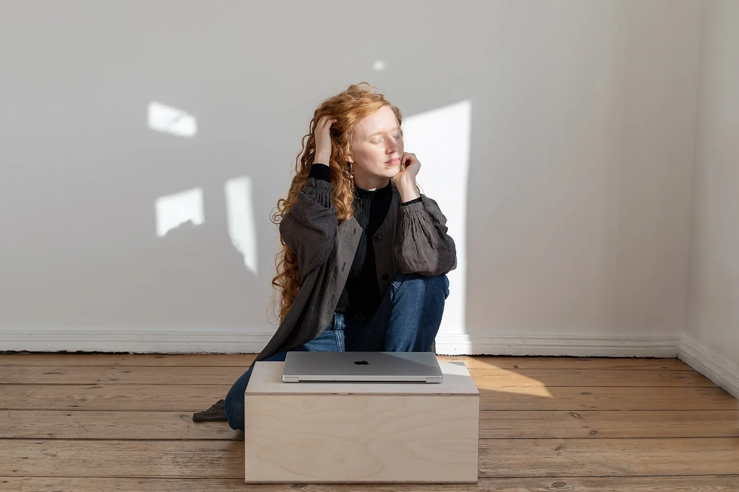 Junge Frau mit langen roten Locken sitzt entspannt auf dem Boden, lehnt den Kopf an die Hand, mit geschlossenen Augen, neben einem Laptop auf einem Holzblock, im Schatten einer Fensterzeichnung, im Raum mit Holzfußboden.