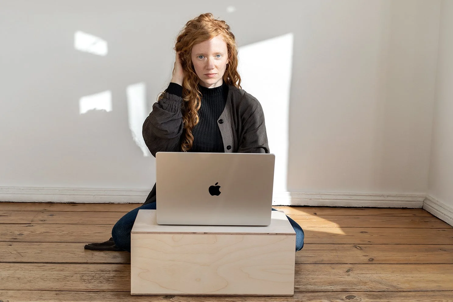 Junge Frau sitzt auf dem Boden, vor einem Laptop, in einem Raum mit Holzboden und weißer Wand, Sonnenlicht wirft Schatten an die Wand.