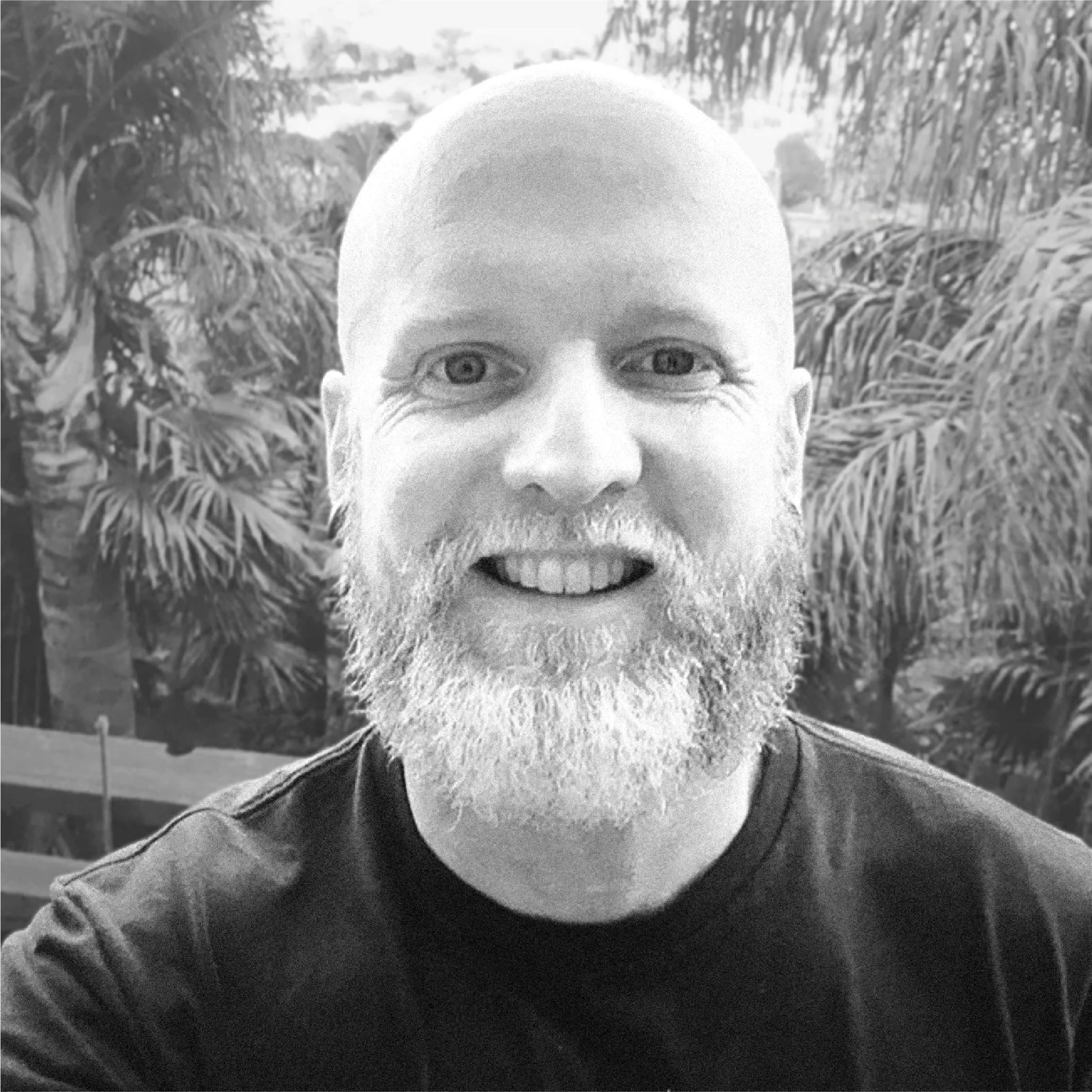 Close-up of a smiling bald man with a background of dense foliage or palm trees.
