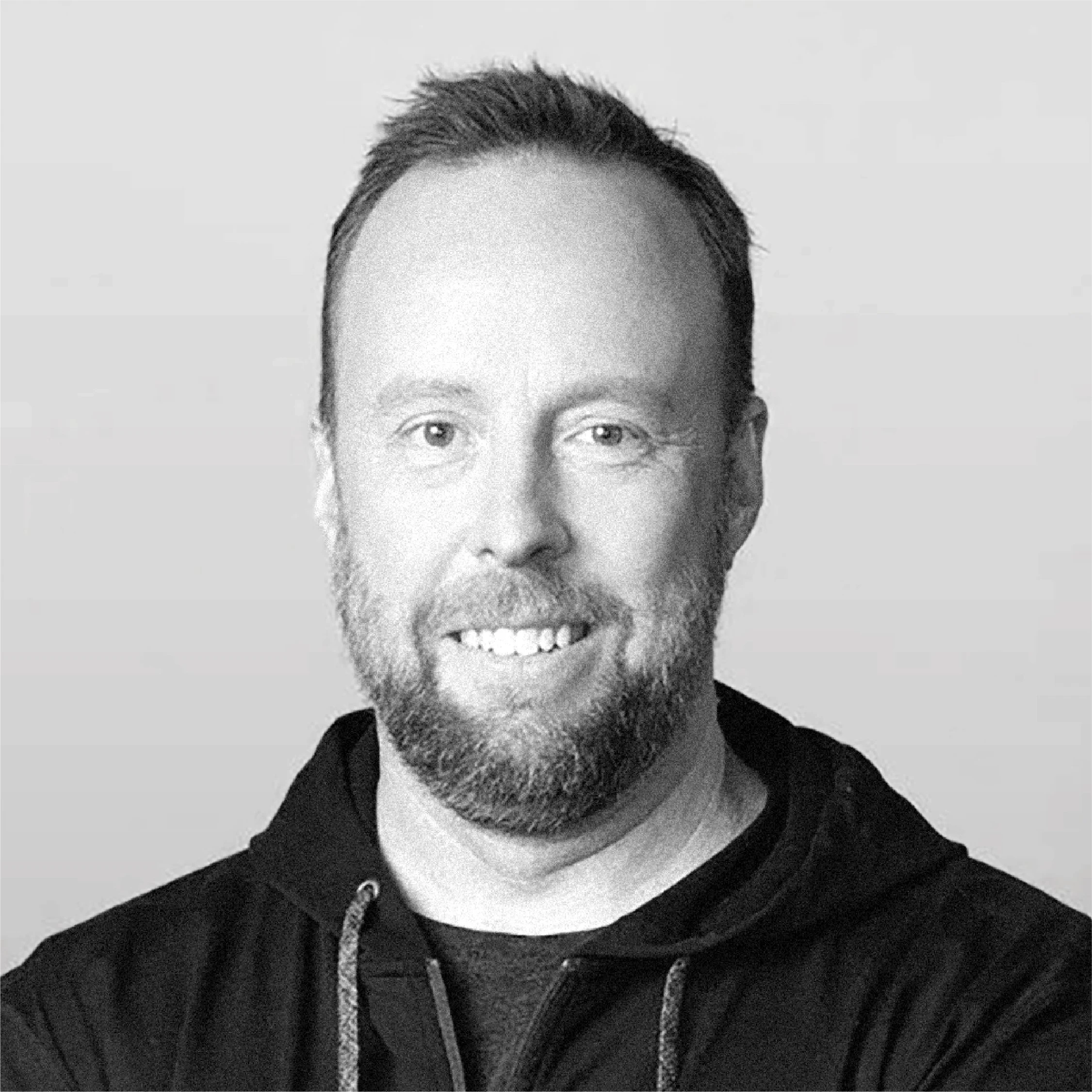 Black and white portrait image of a man with short hair, glasses, a beard, and mustache, smiling and wearing a hoodie.