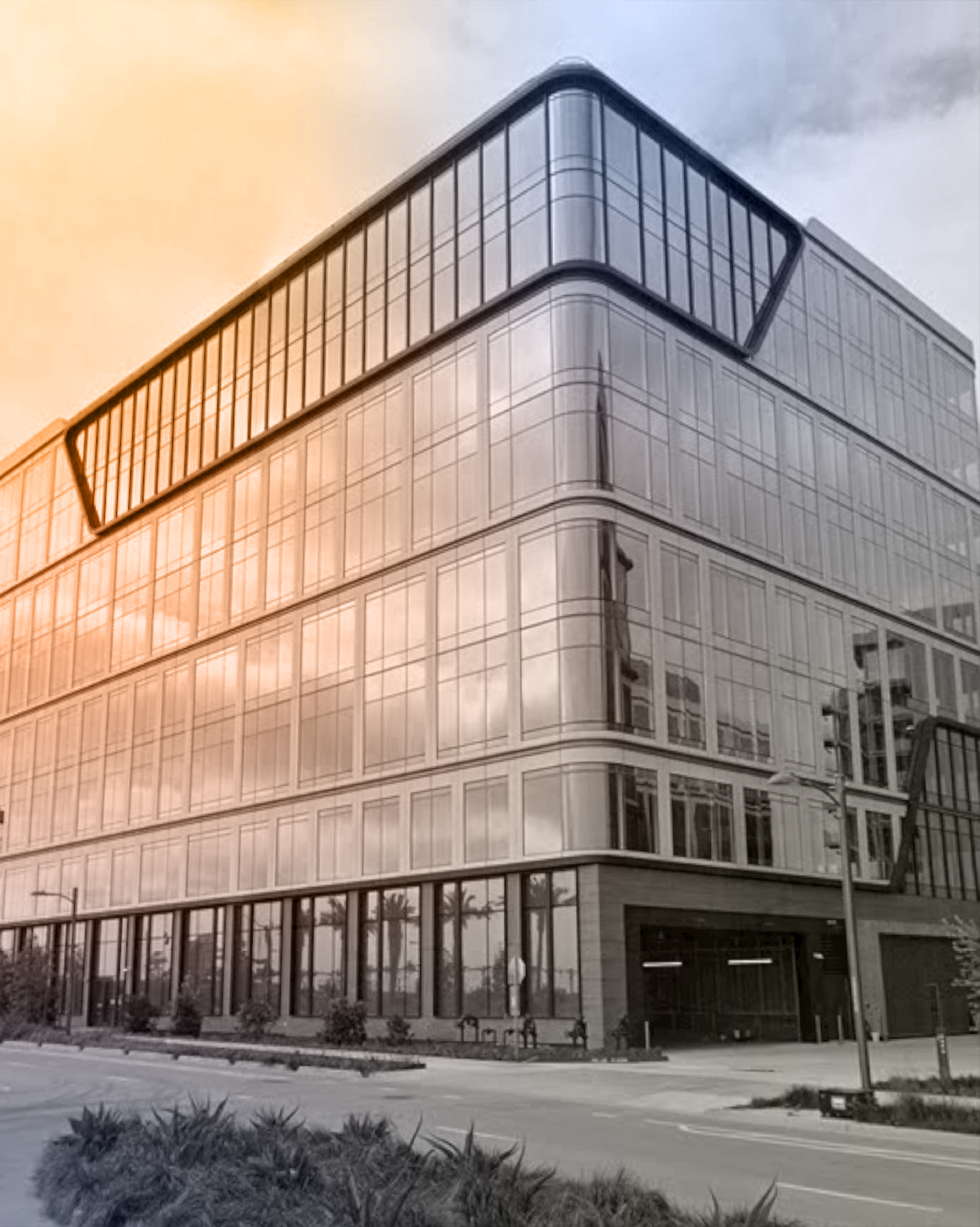 Modern glass office building with rounded corners and multiple floors. The building features reflective windows and is located on a street with landscaping in front. The sky appears overcast.