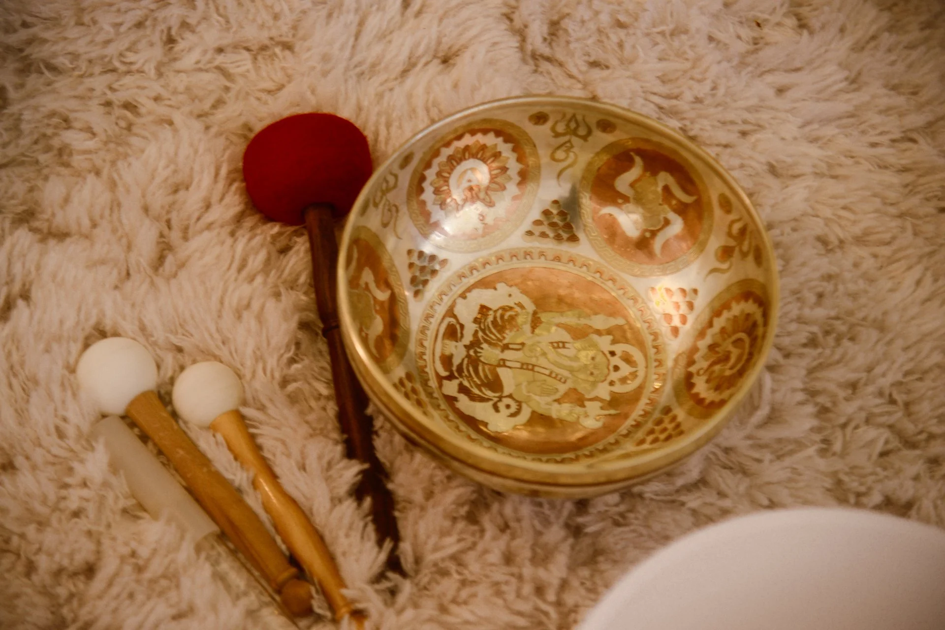 Bol décoratif doré avec motifs traditionnels, deux bâtons de percussion, une balle rouge et deux balles blanches, tous placés sur un tapis en peluche beige.