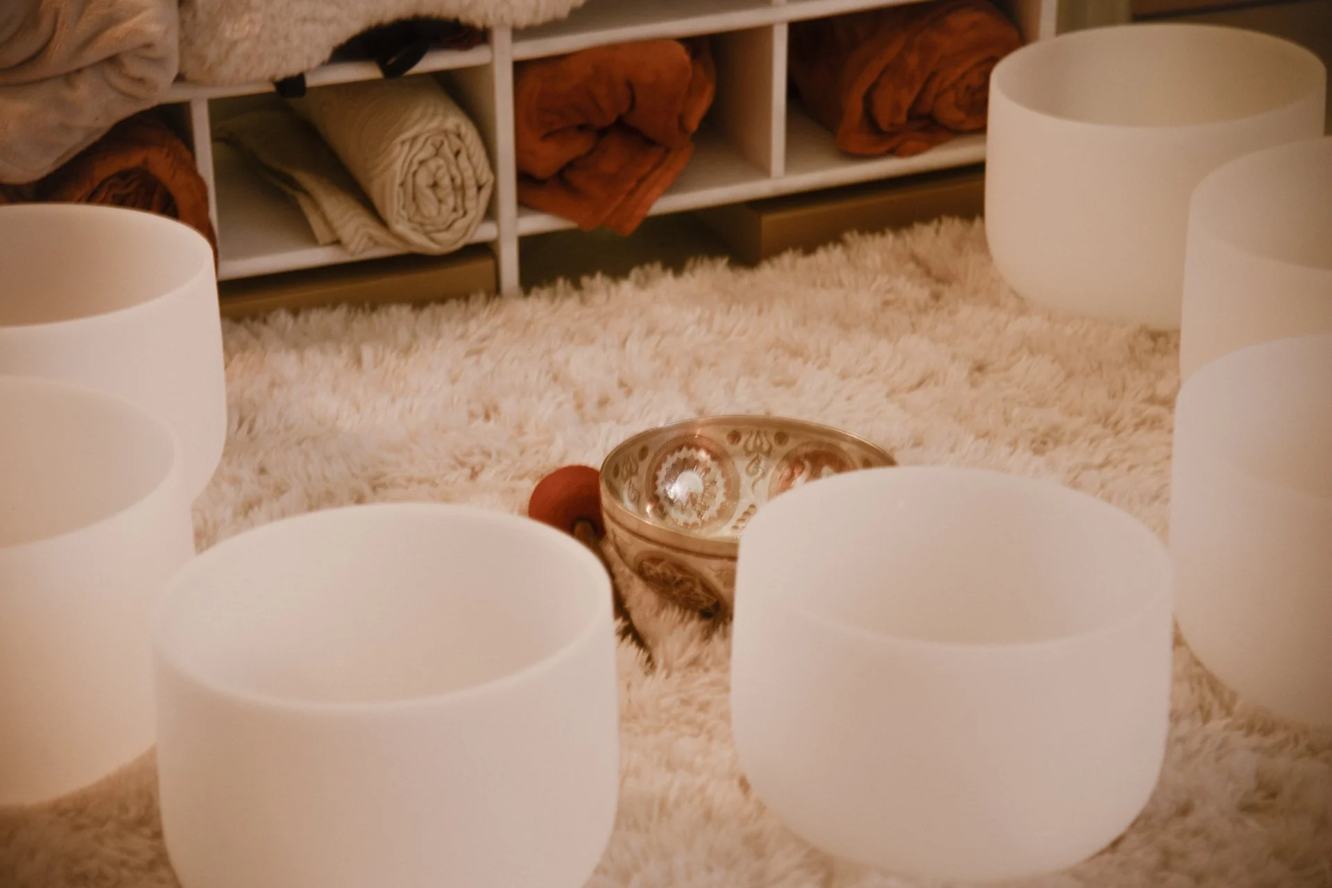 Cinq bols de cristal blancs formant un cercle, placés sur un tapis ou une couverture pelucheuse, avec un bol en céramique ou en métal orné au centre et une petite boule en bois ou pierre à côté. En arrière-plan, un meuble avec des étagères contenant des tissus colorés ou des couvertures roulées.