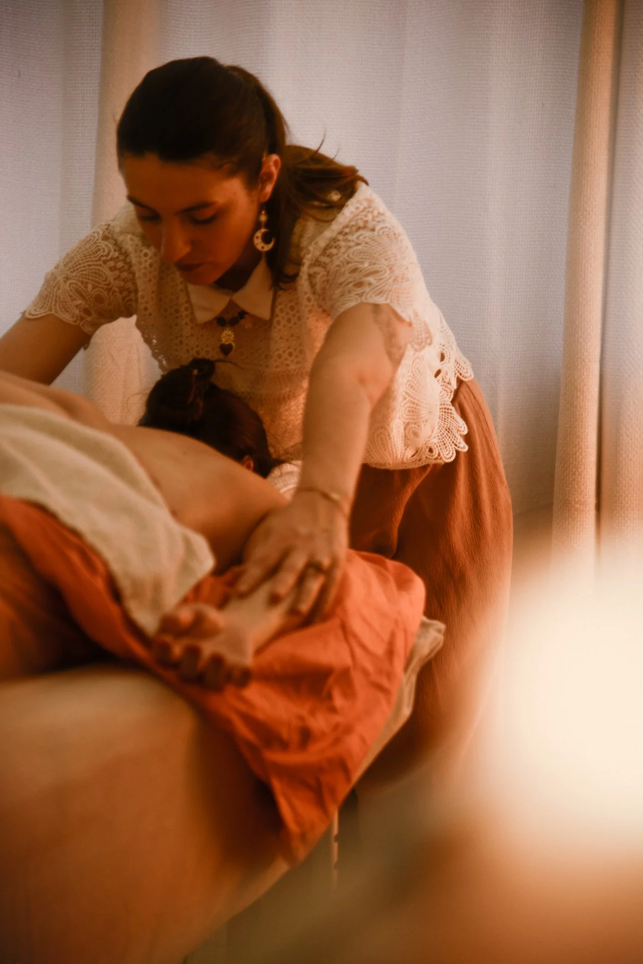 Une femme allongée sur un lit, reçoit un massage de la part d'une autre femme qui porte une t-shirt en dentelle blanc, avec des boucles d'oreilles et un collier.