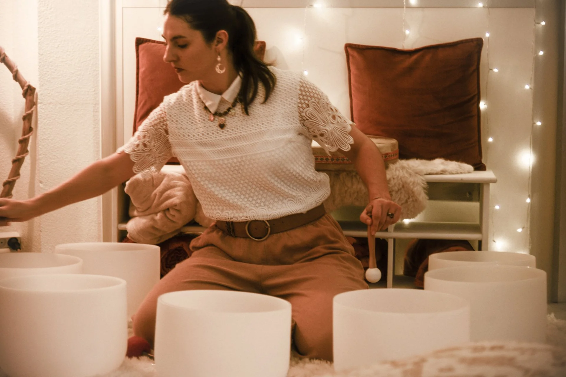 Une femme joue des bols de cristal dans une pièce chaleureuse décorée de guirlandes lumineuses et de coussins.