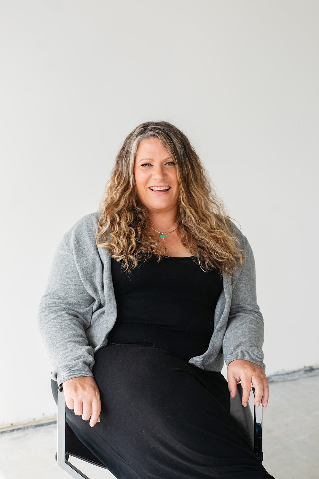 Chantale Spurrier with long curly hair wearing a black top, gray cardigan, and black skirt, sitting on a white chair against a plain white background, smiling at the camera.