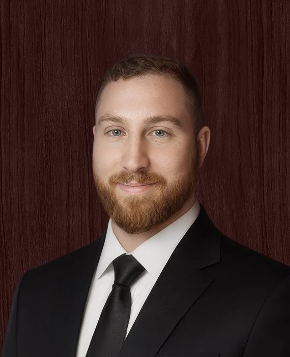 Dan Santucci, Project Manager and Municipal Sector Lead, V2CM, wearing a black suit, white shirt, and black tie, standing in front of a wooden background