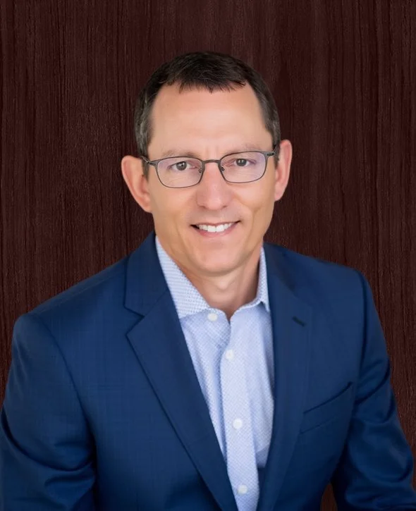 Marius Veldtman, President and CEO, V2CM  in a blue suit with a light blue collared shirt, smiling, seated in front of a wooden background.