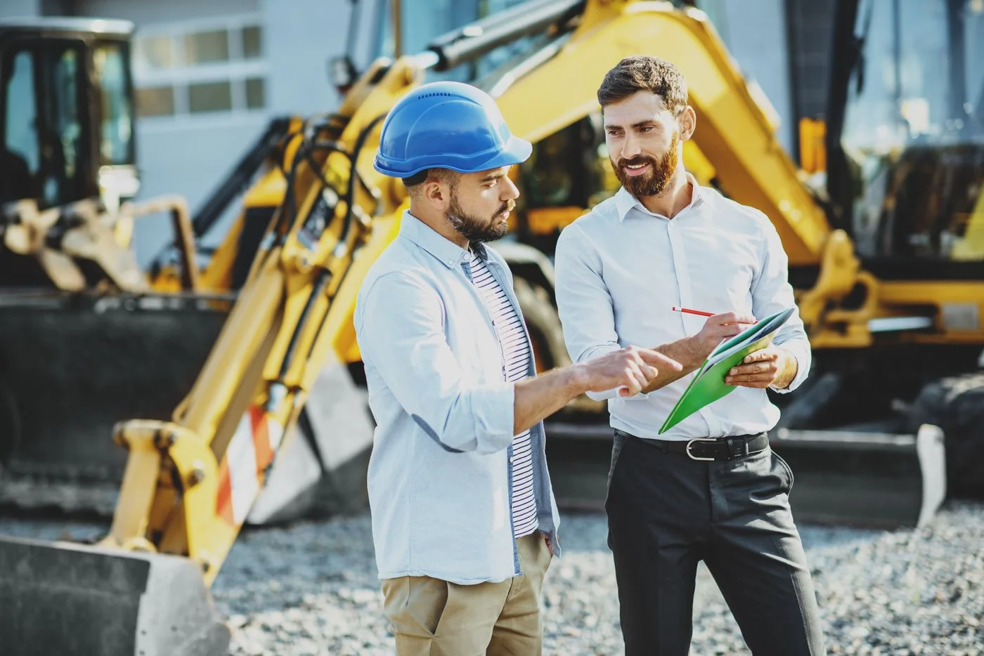 Two men talking at a construction site with heavy machinery in the background, one wearing a blue safety helmet and gloves, the other holding a clipboard and pen.