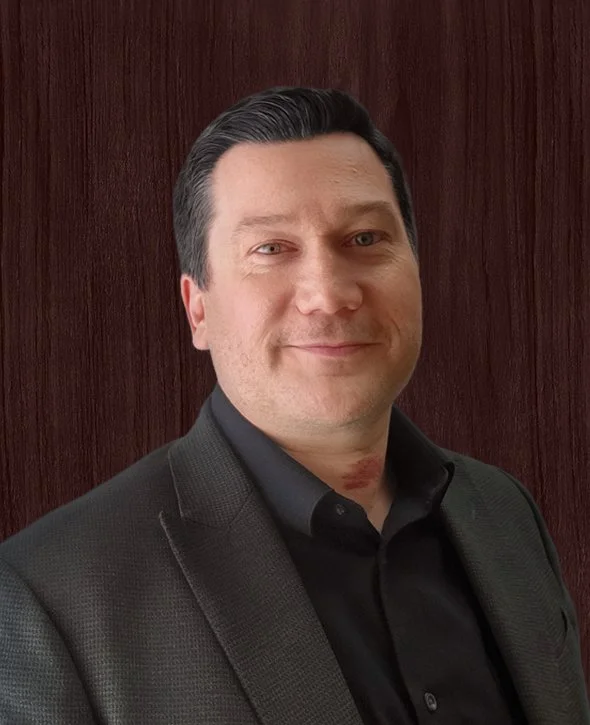 Jared Riehl, Vice President, Finance V2CM, in a dark suit and shirt standing in front of a wooden wall, smiling.