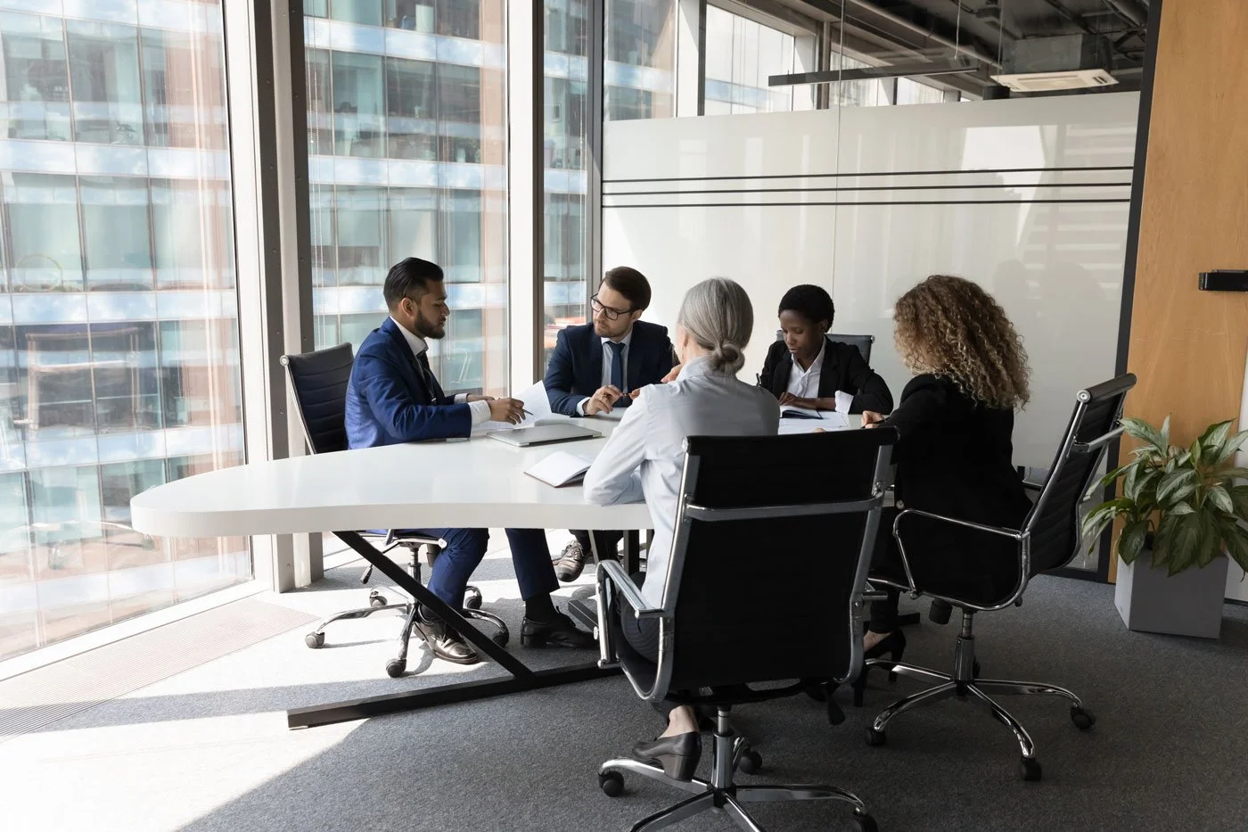 A diverse group of five business professionals, three women and two men, are seated around a white conference table in a modern office with large windows. They appear to be engaged in a discussion, with documents and notebooks on the table. A potted plant is visible in the corner of the room.