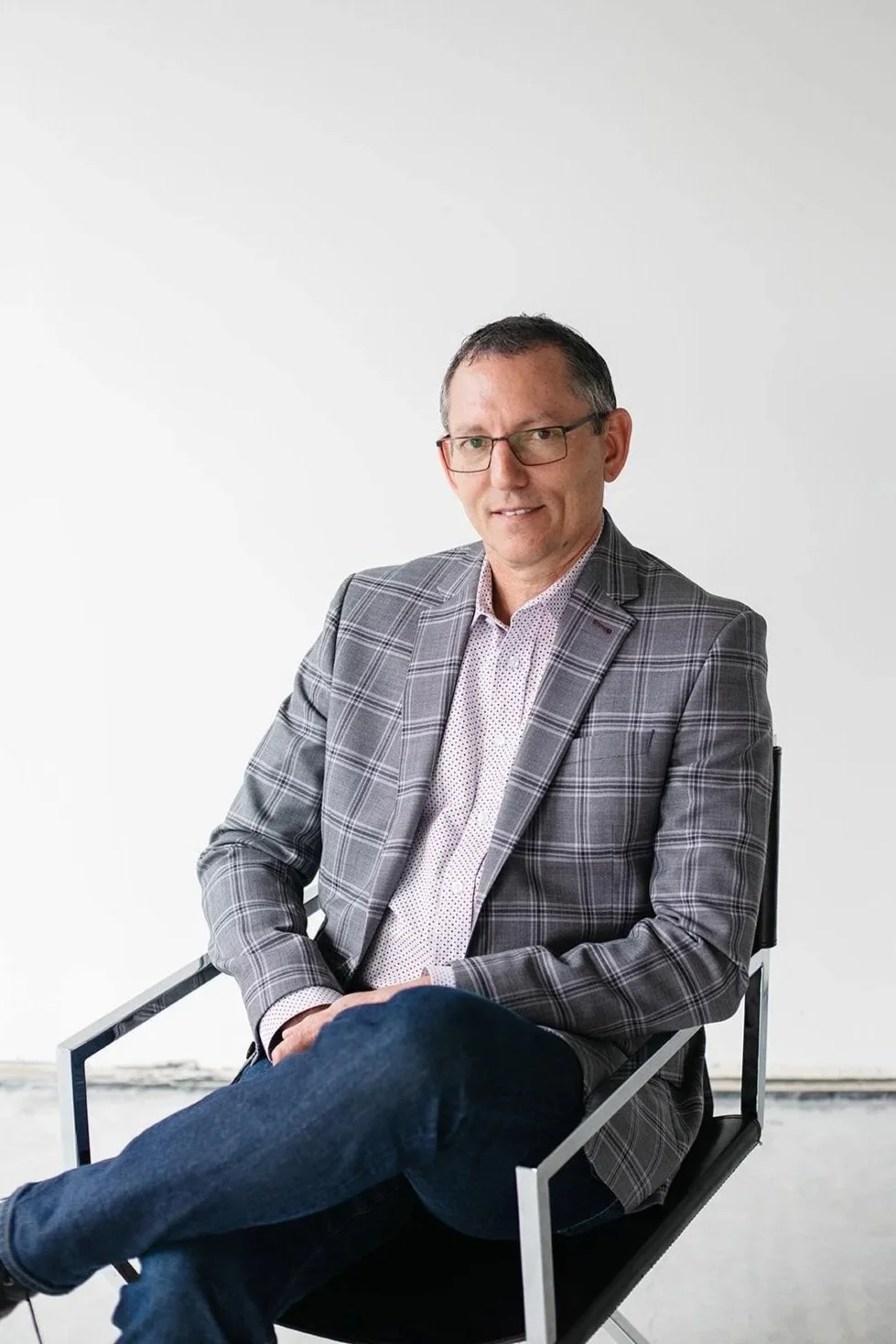 Marius Veldtman, President and CEO, V2CM in a blue suit with a light blue collared shirt, smiling, seated in front of a wooden background.