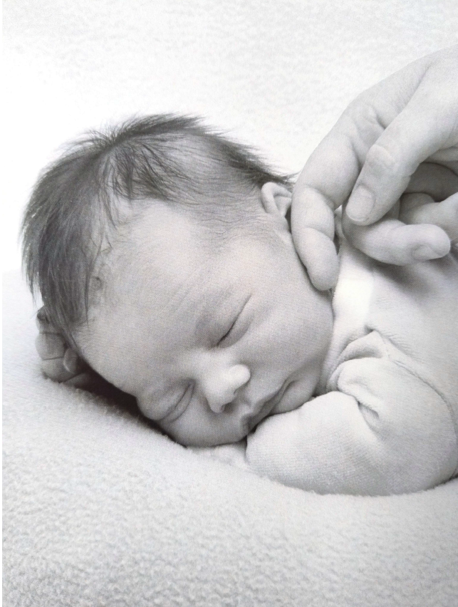 Un bébé endormi, avec une main d'adulte touchant son visage, en noir et blanc.