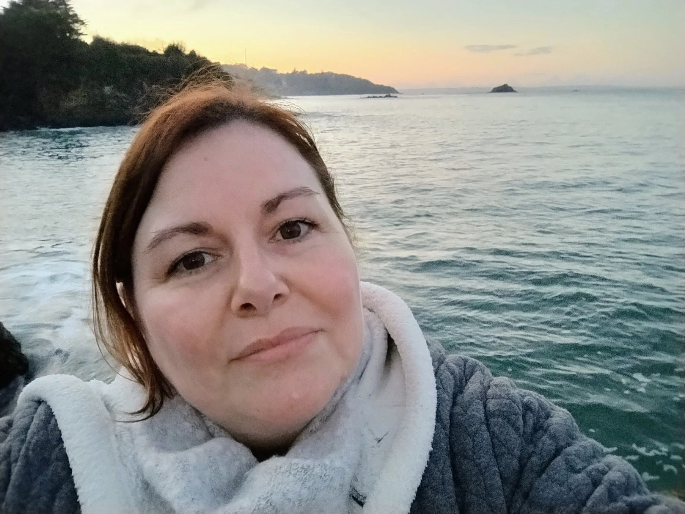 Une femme souriante avec des cheveux bruns, portant une veste grise avec intérieur blanc, se trouve près d'une mer calme lors du coucher du soleil, avec des îles en arrière-plan.