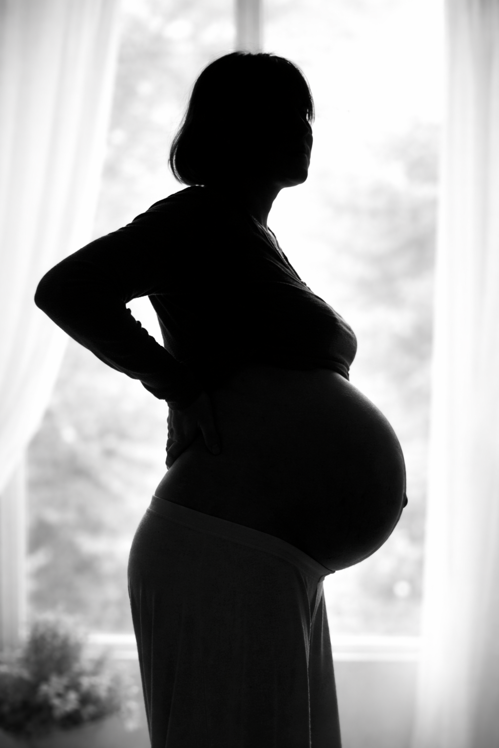 Silhouette d'une femme enceinte, vue de profil, debout devant une fenêtre avec des rideaux légers.