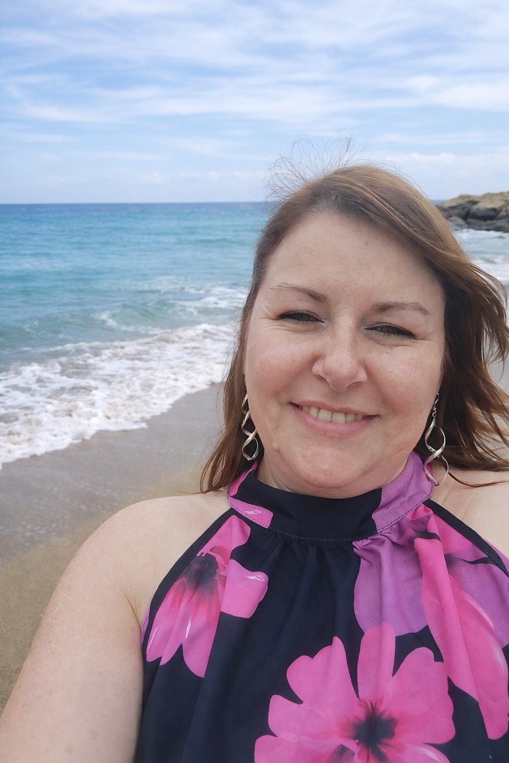 Une femme souriante à la plage, avec la mer et le ciel en arrière-plan, portant une robe à fleurs roses et violettes.