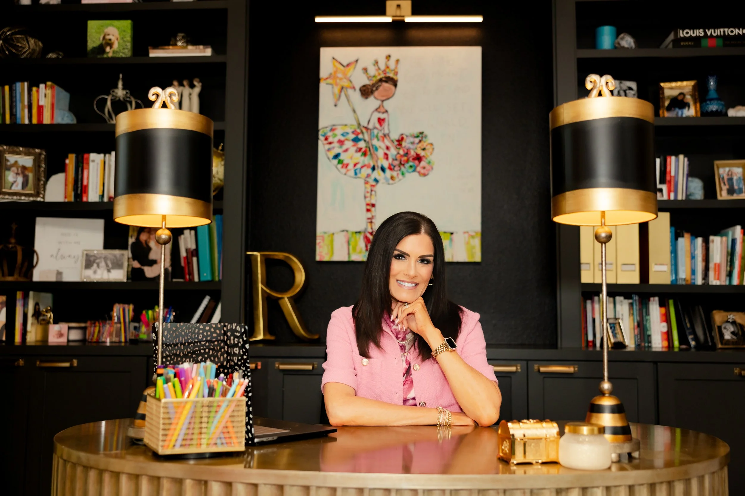 A woman with long dark hair and a pink blazer sitting at a wooden table in a well-decorated room with bookshelves, artwork, and two black and gold lamps.