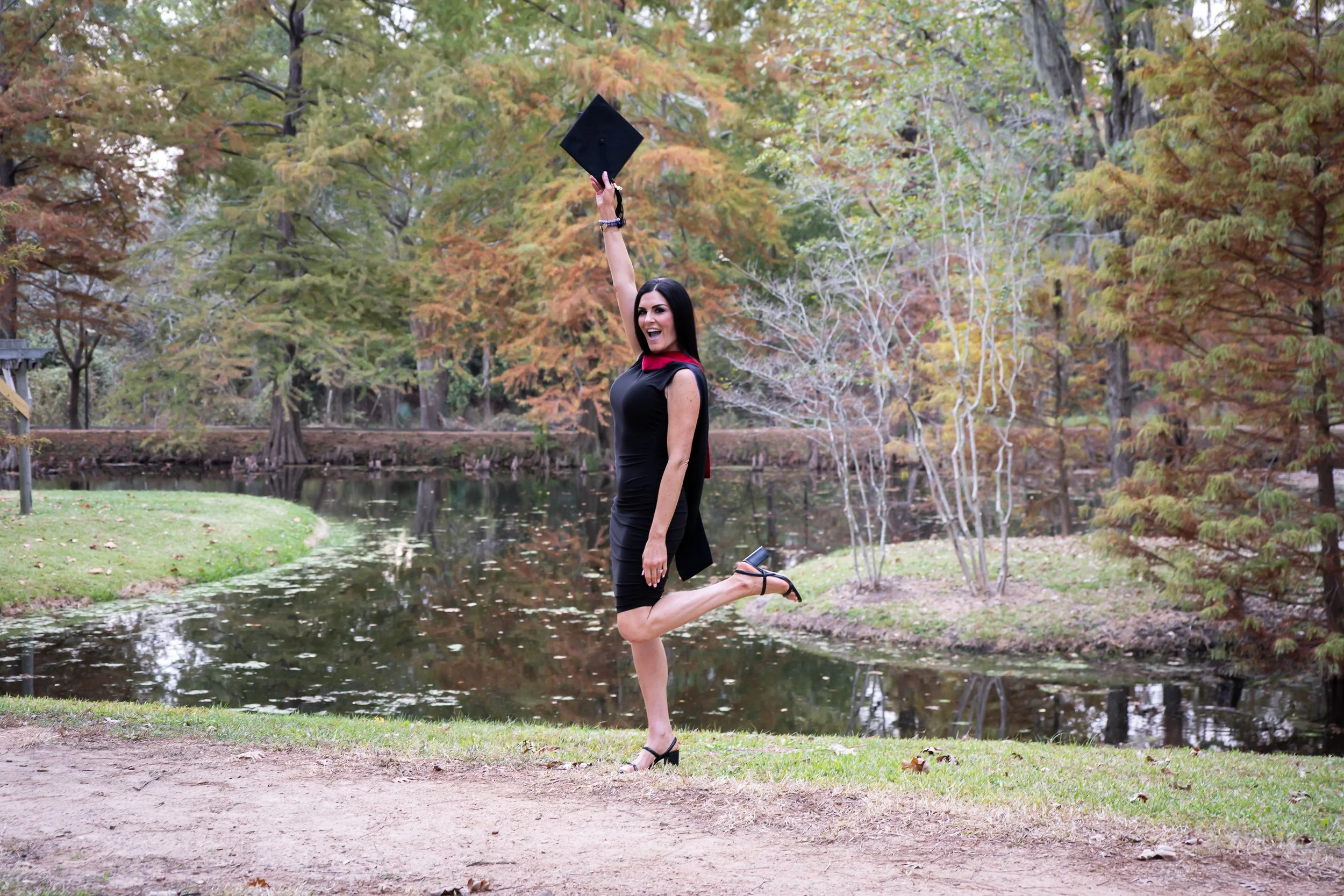 A woman in a black dress and heels joyfully jumping with one knee raised and holding a graduation cap in the air near a pond surrounded by autumn trees.