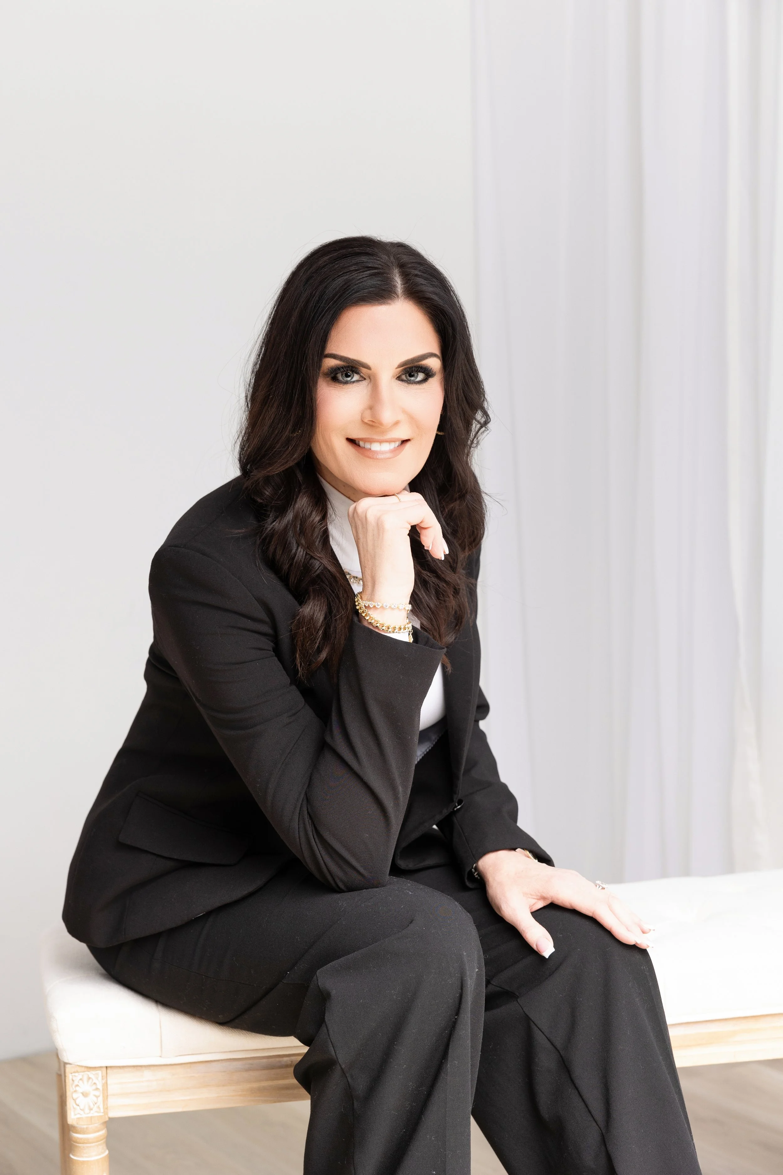 A woman with long dark hair, dressed in a black blazer and pants, sitting on a white bench, smiling and resting her chin on her hand.