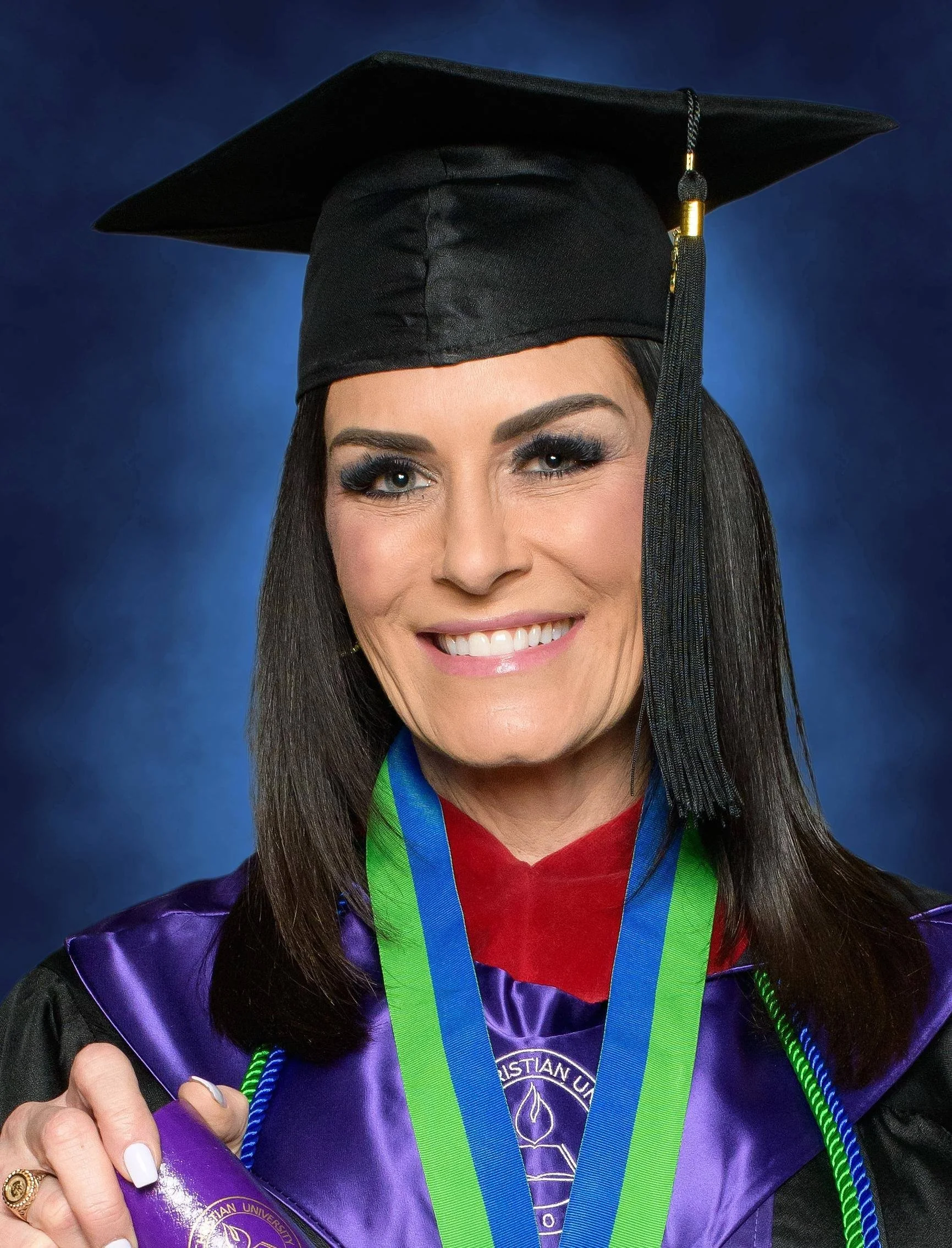 A woman with black hair and makeup, wearing a black graduation cap and gown with colorful accents, smiling and holding a diploma with a green and blue ribbon, against a blue background.