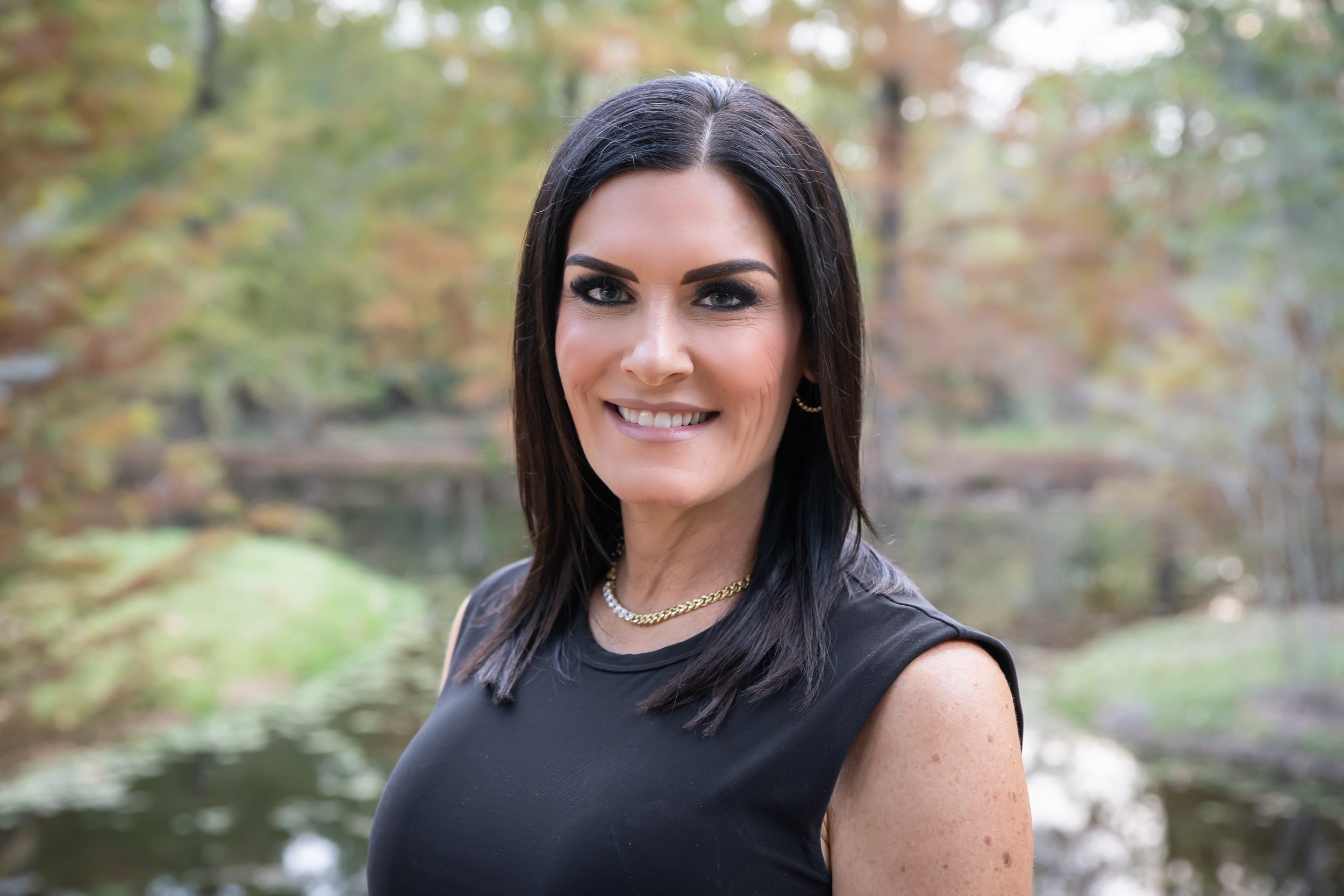 A woman with dark hair and makeup smiling outdoors in front of a blurred background of trees and a body of water.