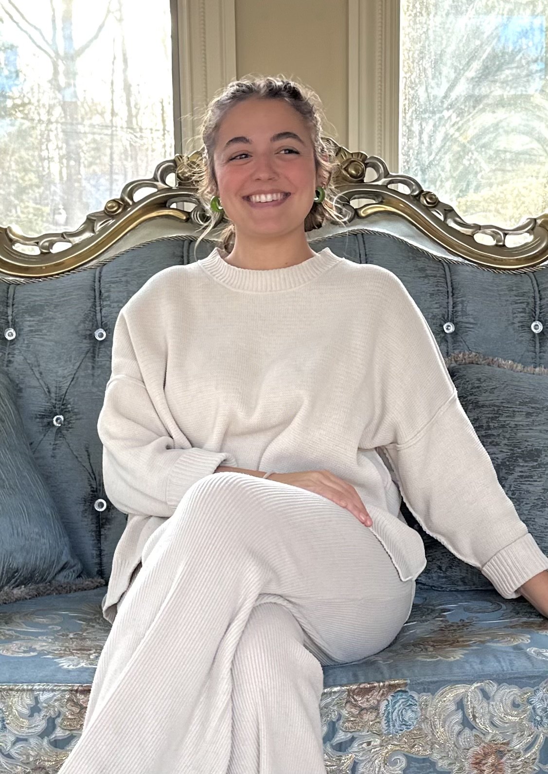 A woman with curly hair smiling, sitting on an ornate vintage-style blue sofa with gold trim, in front of a large window with trees outside, wearing a cream-colored sweatshirt and pants, green earrings.