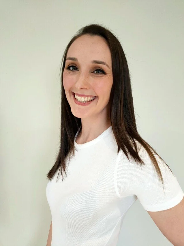 A young woman with long dark hair smiling and winking at the camera, wearing a white t-shirt against a plain light-colored background.