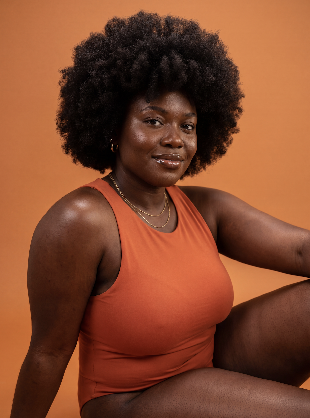 Portrait of a Black woman with natural curly hair, wearing an orange sleeveless top, gold jewelry, and sitting against a warm orange background.