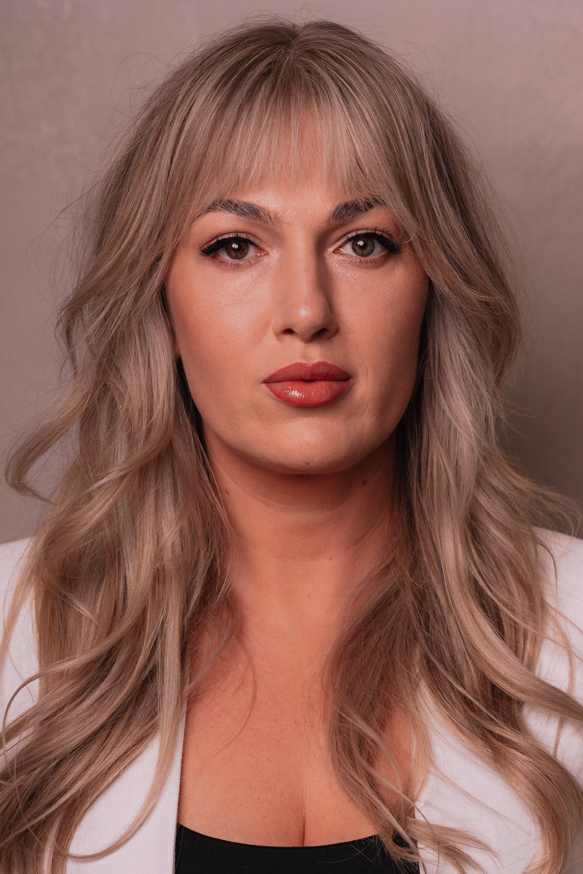 Portret van een vrouw met lang, golvend, lichtbruin haar en make-up, gekleed in een witte jas en een zwarte top.