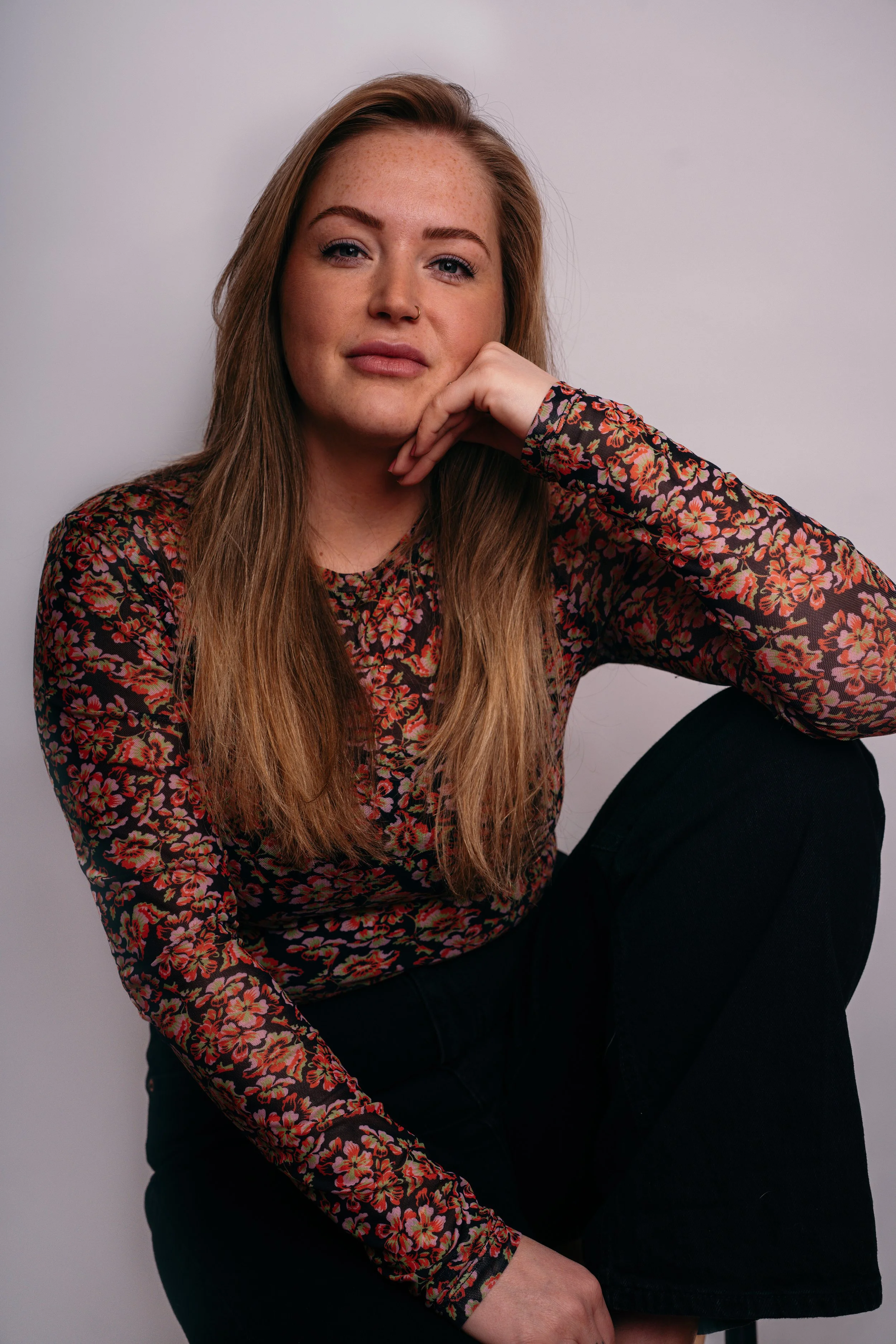 Portret van een jonge vrouw met lang, lichtbruin haar, lichte huid en sproeten, zittend tegen een witte achtergrond, met haar rechterelleboog op haar knie en haar hand onder haar kin, gekleed in een bloemenprint top en zwarte broek.