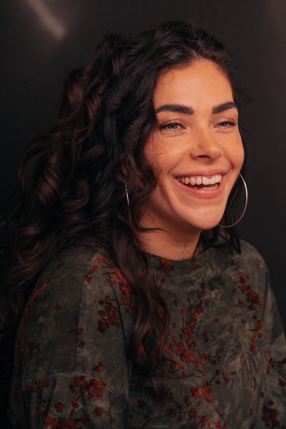 Close-up van een lachende vrouw met zwarte, gekrulde haar en grote oorbellen, tegen een donkere achtergrond.