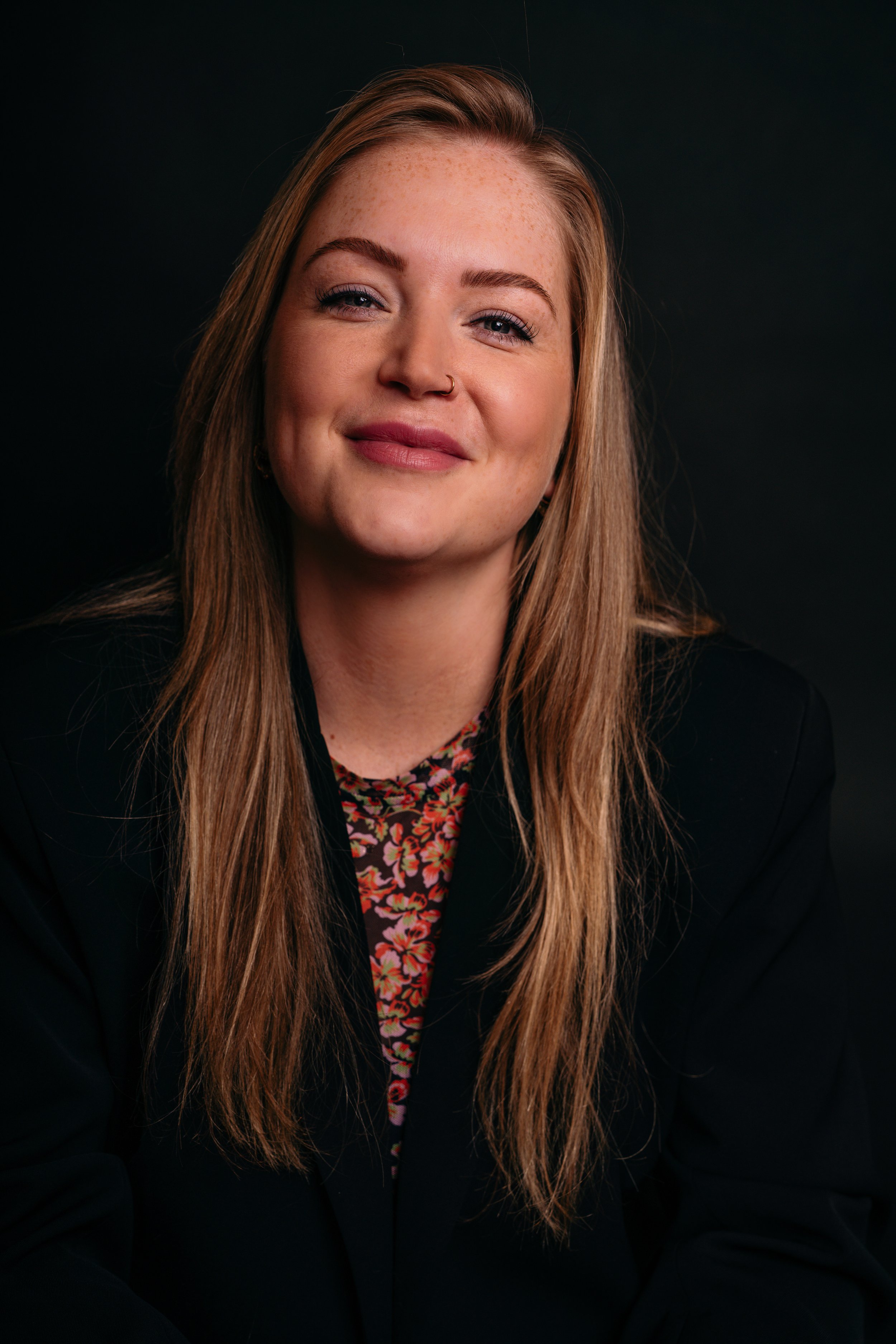 Portret van een lachende jonge vrouw met lang, lichtbruin haar, een neuslingering en een zwarte achtergrond.
