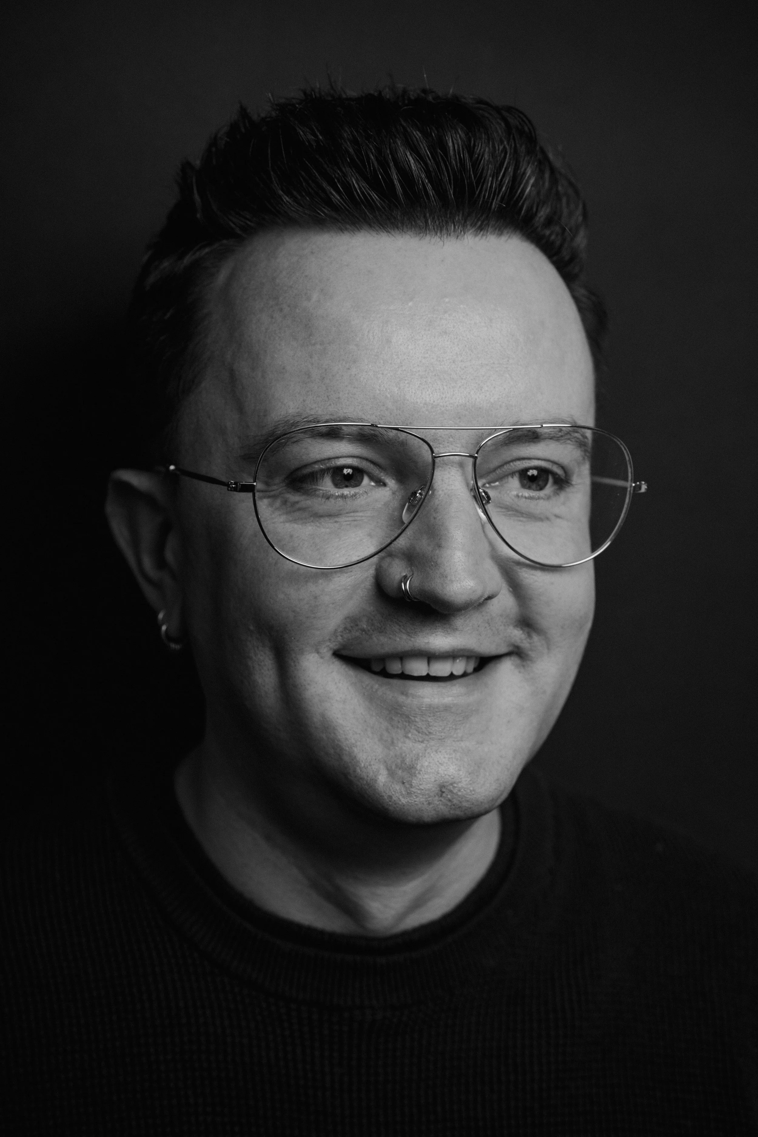 Zwart-witte foto van een persoon met kort haar, grote ronde bril, neusring en kleine oorbellen, lachend met tanden zichtbaar.