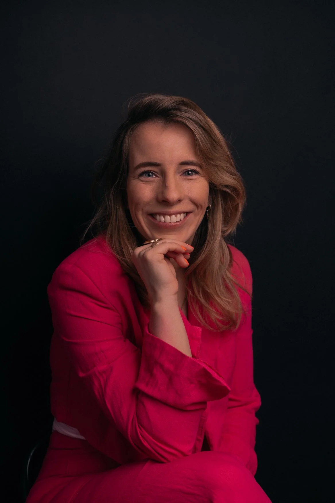 Portret van een lachende vrouw met bruin haar in een roze blouse voor een zwarte achtergrond.