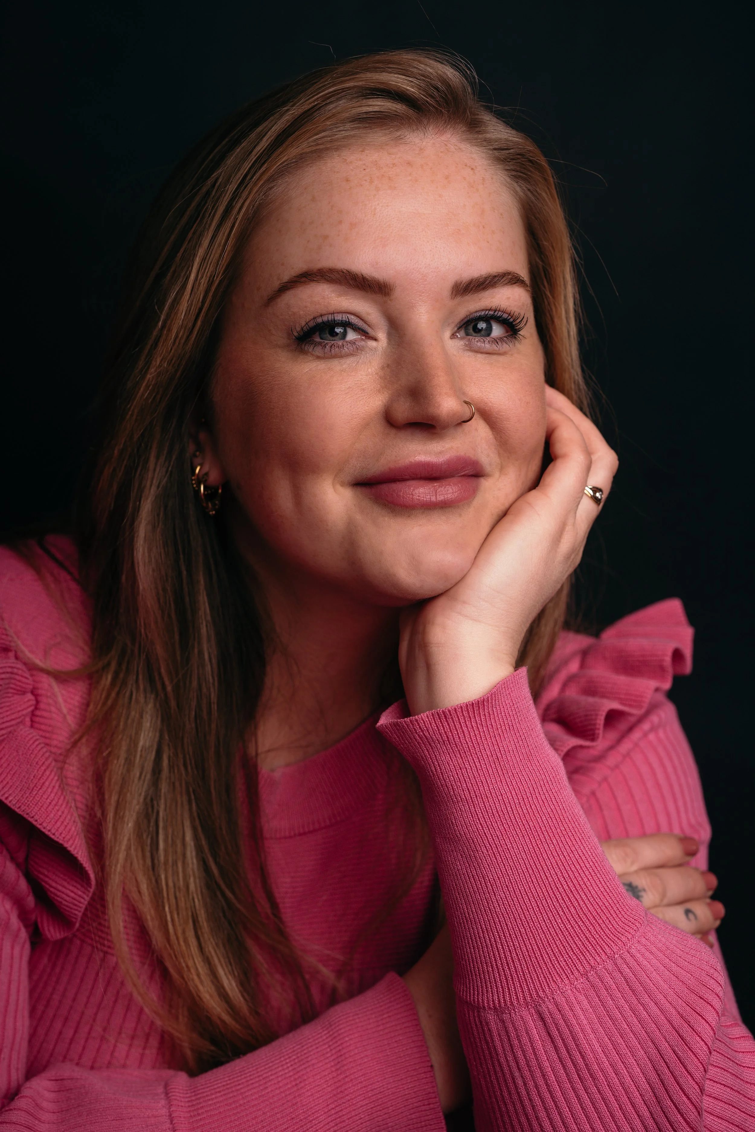 Portret van een vrouw met lang bruin haar, lichte huid met sproeten, blauwe ogen, draag een roze trui en heeft sieraden, glimlacht zacht tegen een zwarte achtergrond.