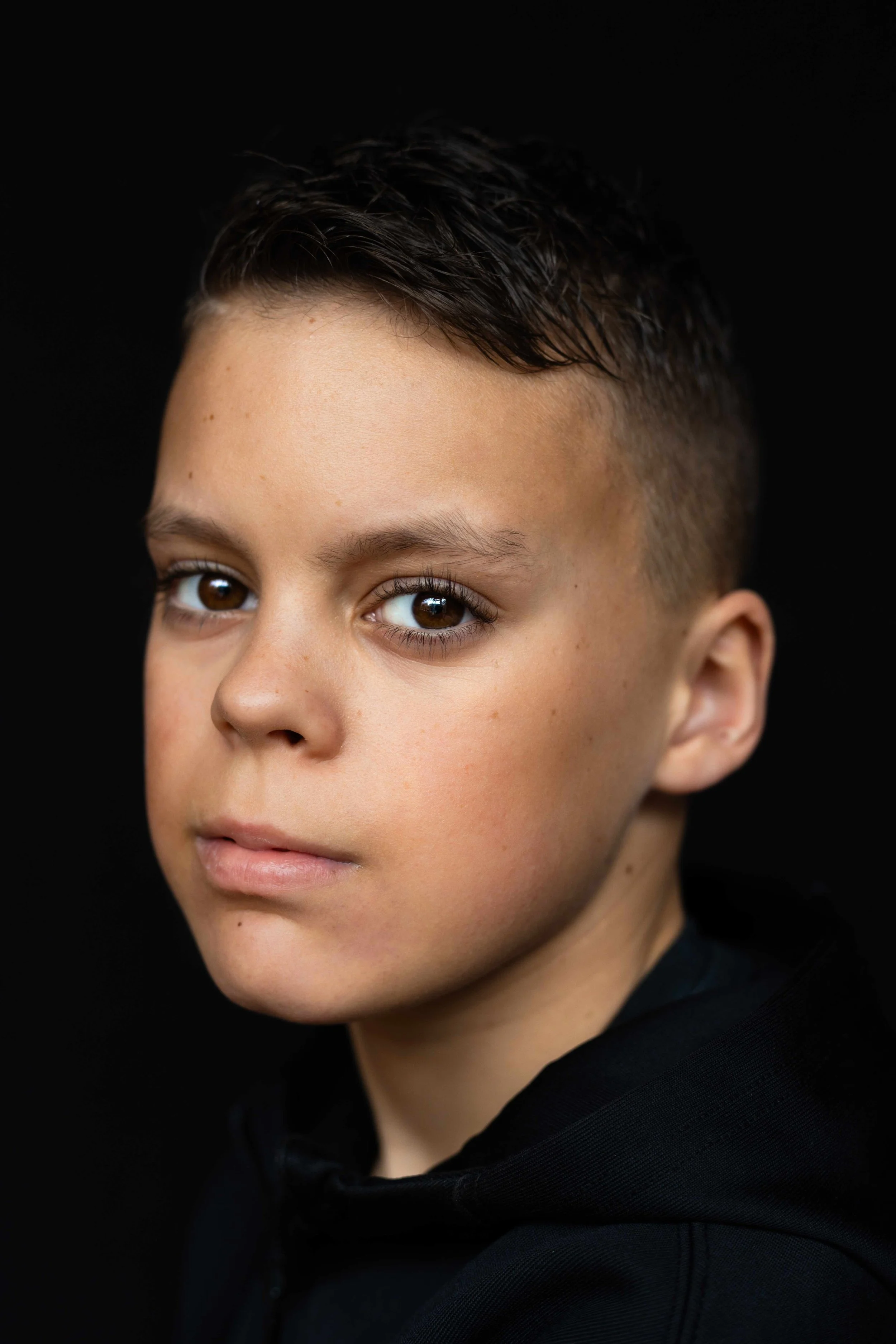 Close-up portret van een jonge jongen met kort, donker haar en bruine ogen, gekleed in een zwart hoodie, zwart achtergrond.