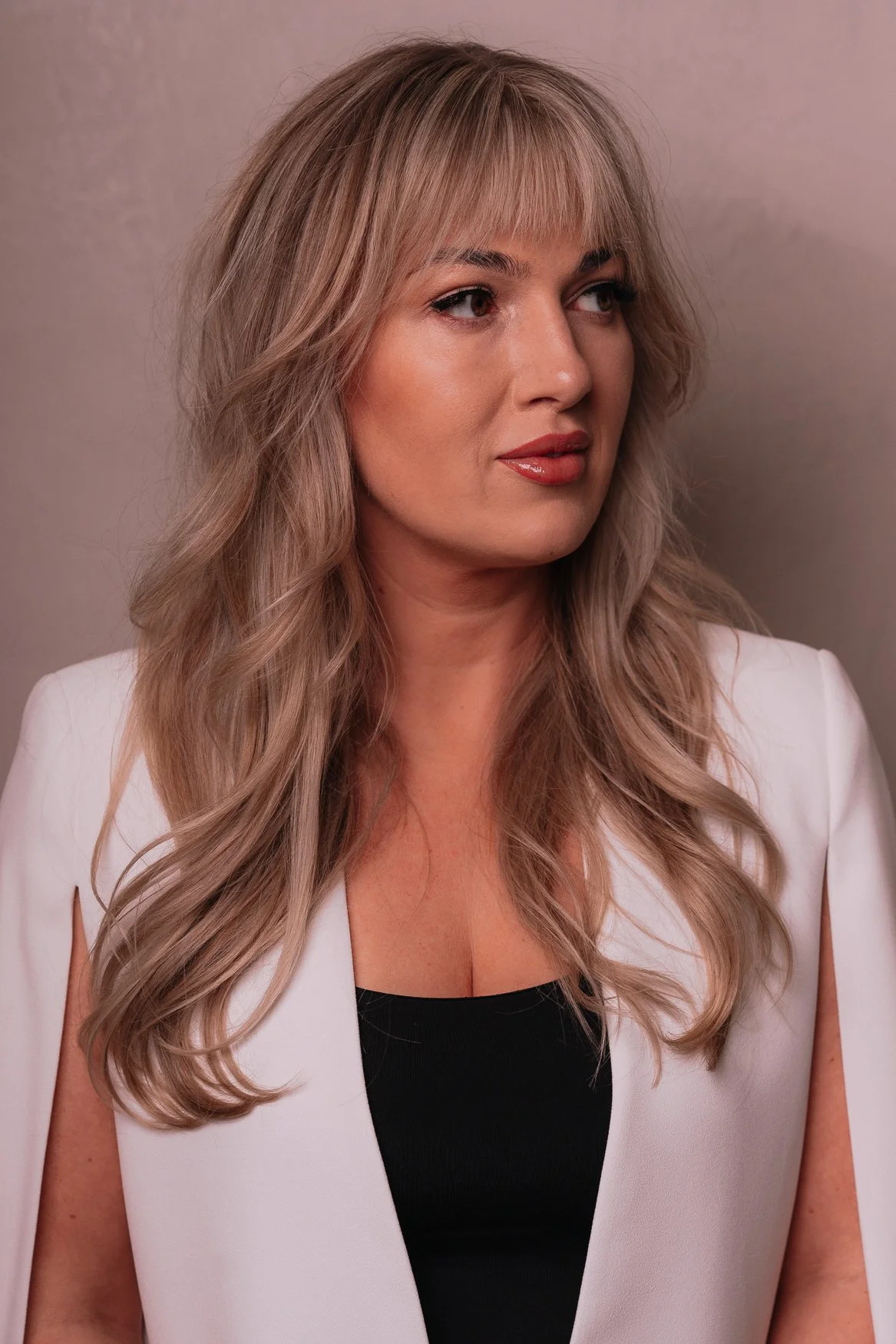 Portret van een vrouw met lange, golvende, blond haar en lichte huid, gekleed in een witte jas en een zwarte top, tegen een effen achtergrond.