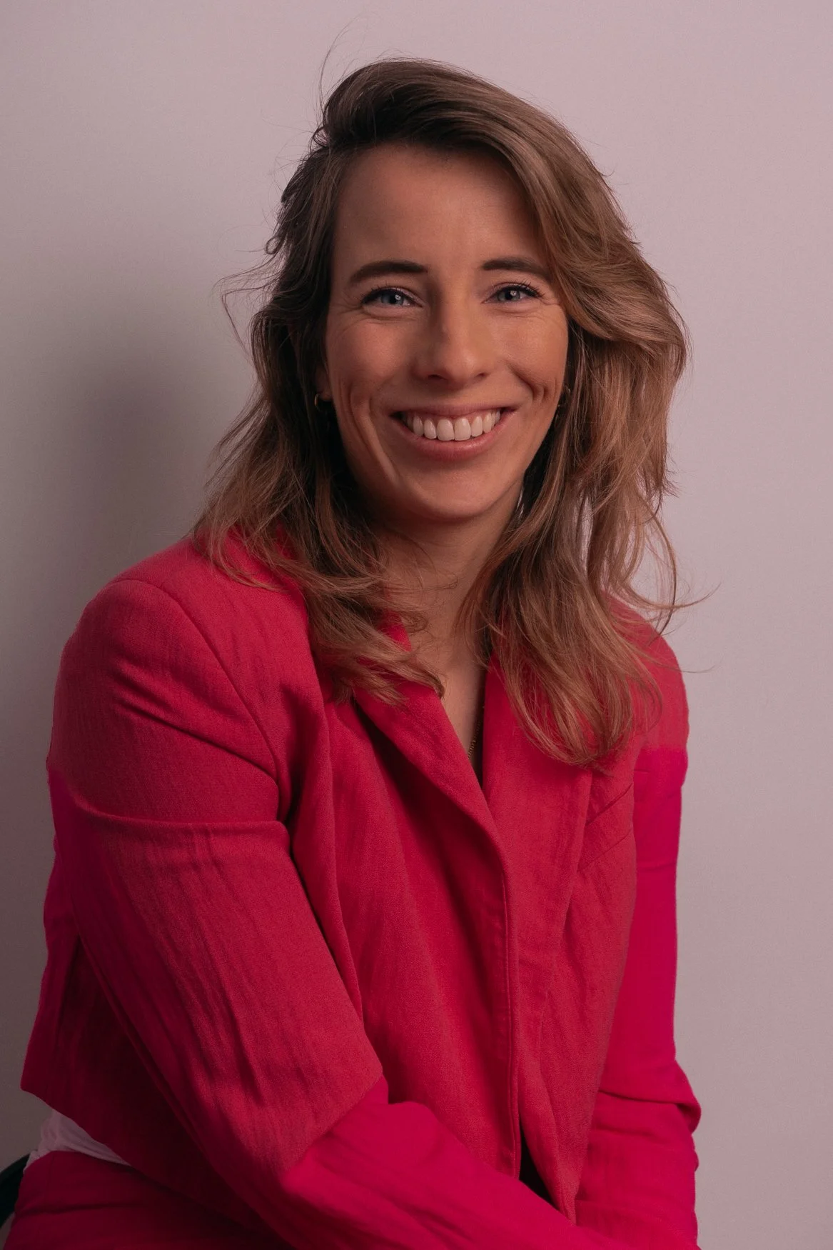 Portret van een lachende vrouw met blond haar, gekleed in een roze jas, tegen een witte achtergrond.