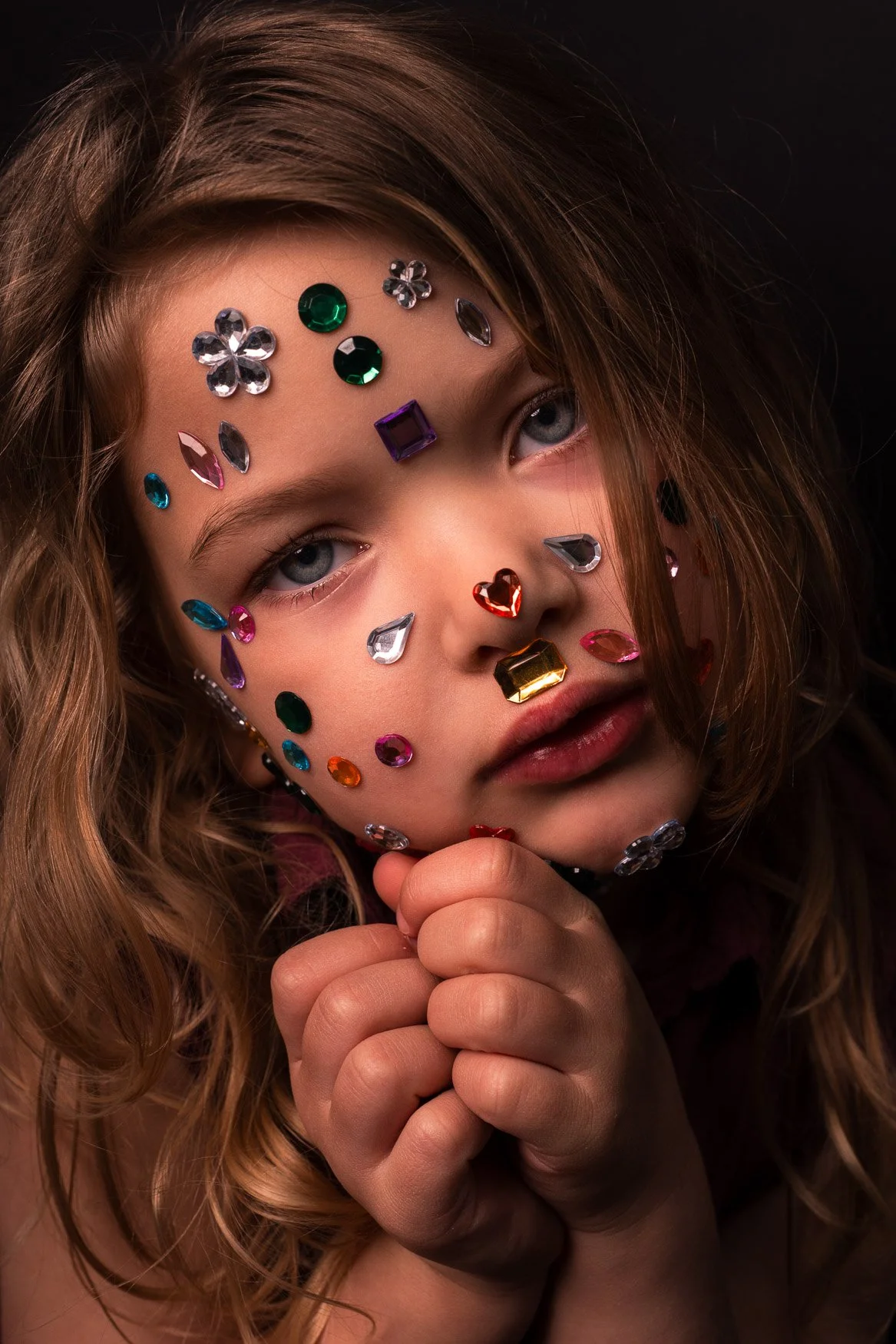 Een close-up van een jonge dochter met lippetal en gezicht bedekt met gekleurde ruitvormige en ronde juwelen, staart dromerig, handen bij haar kin.
