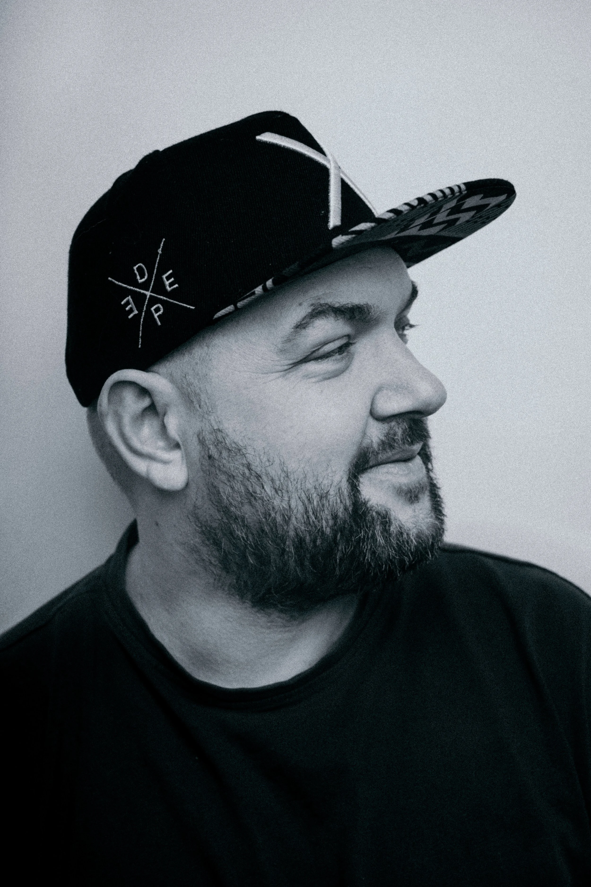 Zwart-wit foto van een man met een baard, die kijkt naar rechts, draagt een zwart T-shirt en een zwarte baseballpet met een grafisch ontwerp op de rand.