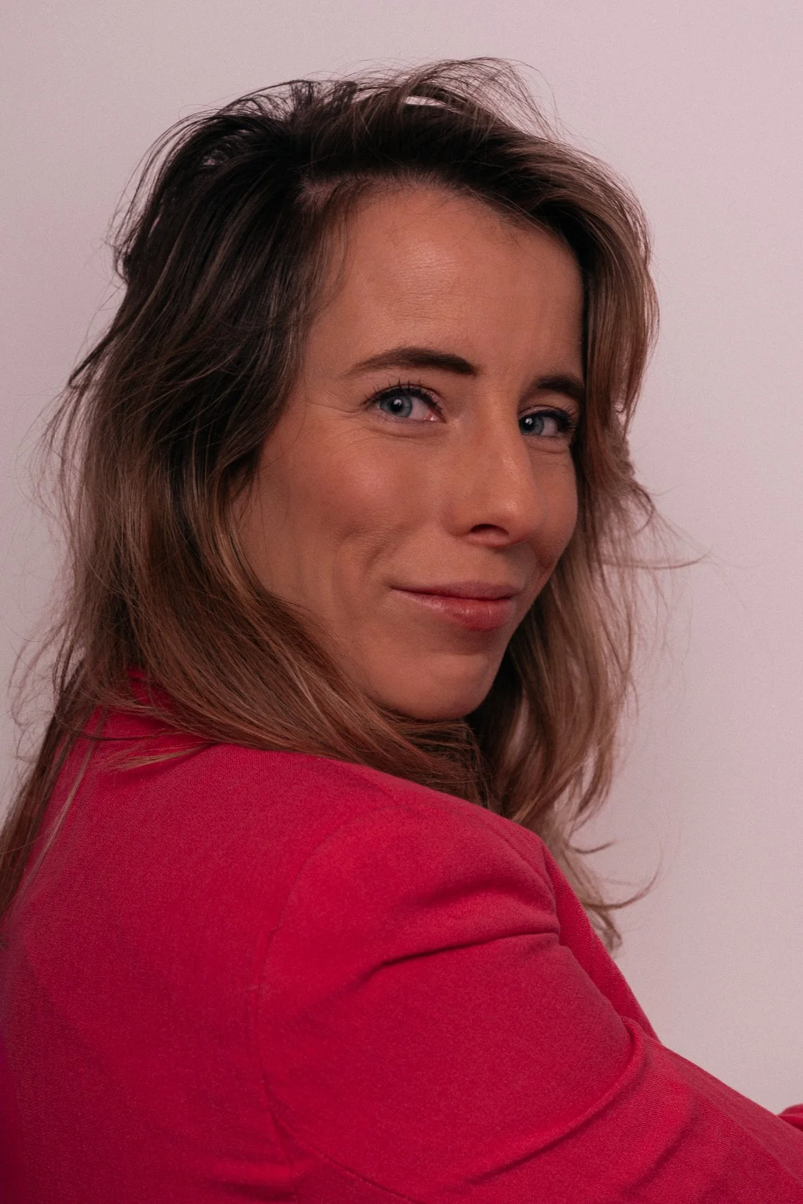 Portret van een vrouw met bruin haar en blauwe ogen die glimlacht, draag een rode jas, op een lichte achtergrond.