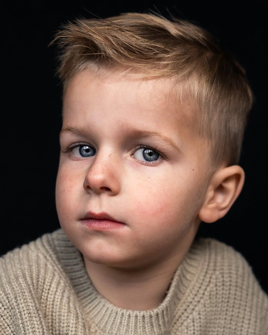 Close-up van een jongen met blauwe ogen, lichtbruin haar en een beige gebreide trui tegen een zwarte achtergrond.