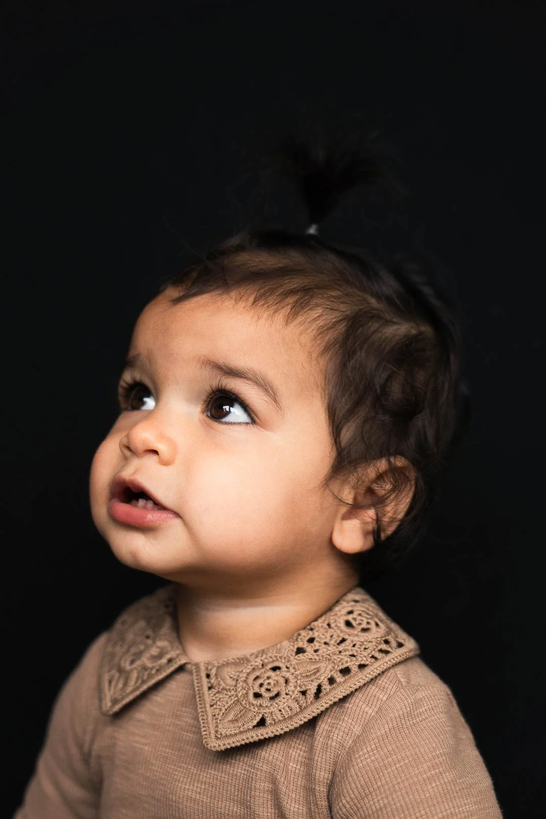 Achtergrondloze foto van een jonge baby met donker haar en grote donkere ogen, gekleed in een beige kledingstuk met een gedetailleerde borduursel op de kraag.